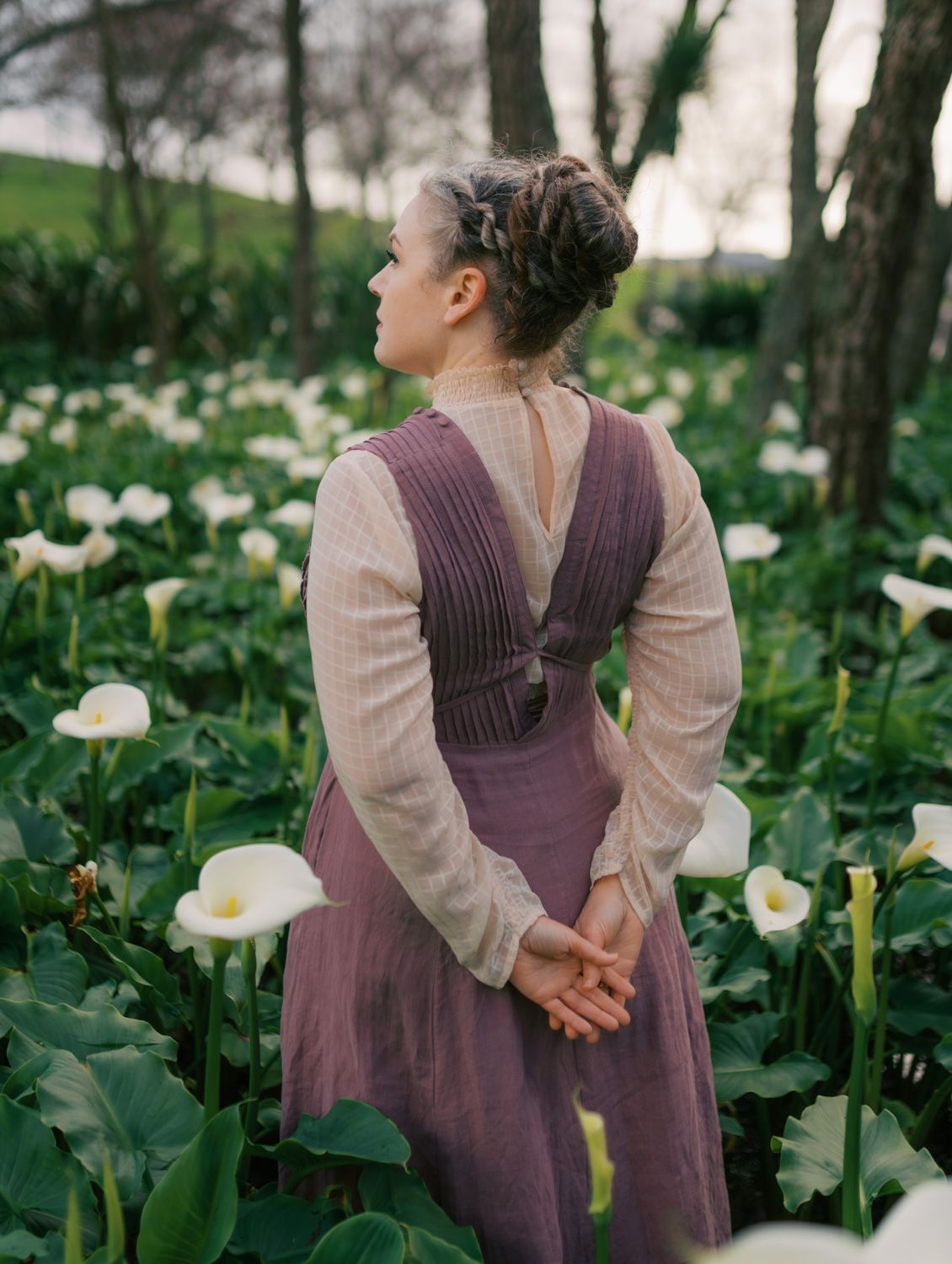 Woman in purple pinafore dress stands among white calla lilies in a lush garden, viewed from behind.