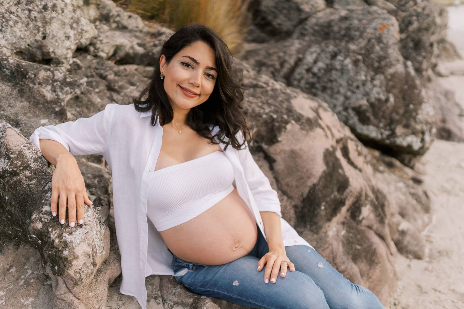 Pregnant woman in white crop top and jeans sitting on rocks at beach, smiling warmly at camera.