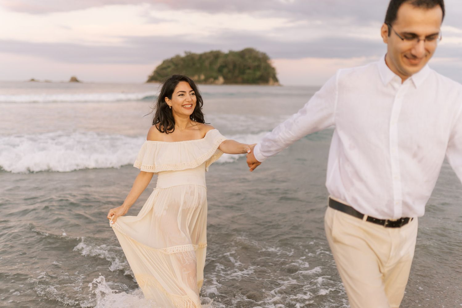 Couple holding hands walking on a beach at sunset, woman in white dress, man in white shirt and khaki pants.