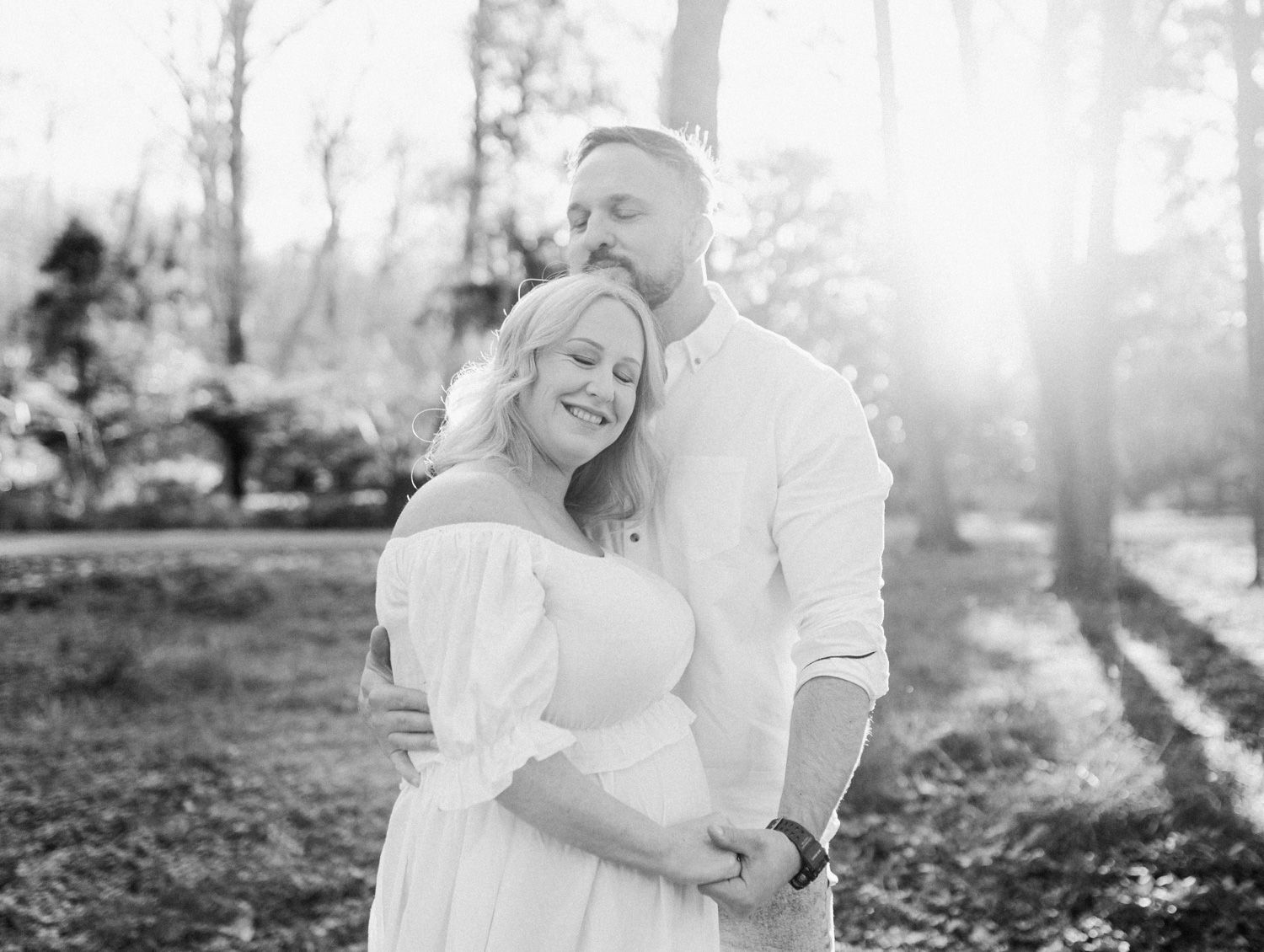 Black and white photo of smiling pregnant couple embracing outdoors, woman in off-shoulder white maternity dress.