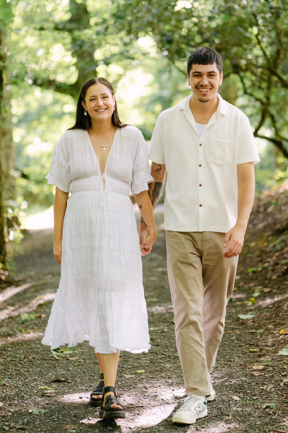 Happy couple walking hand in hand on a shaded forest path, woman in white dress, man in white shirt and khakis.