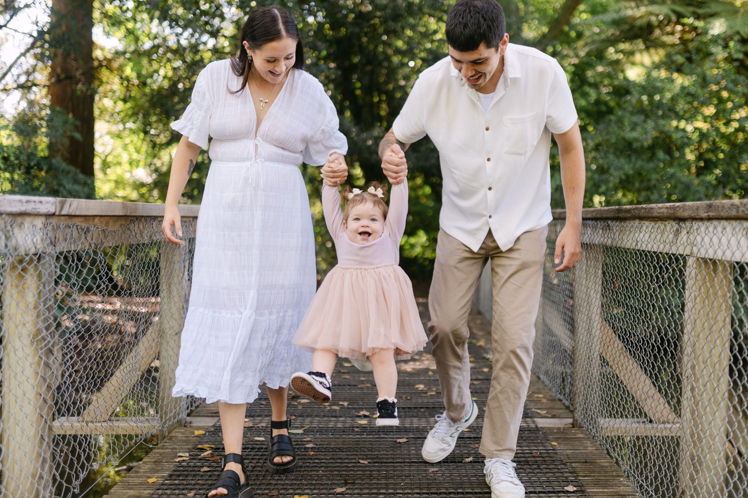 Happy family with young toddler girl holding hands on a wooden bridge surrounded by lush green trees.