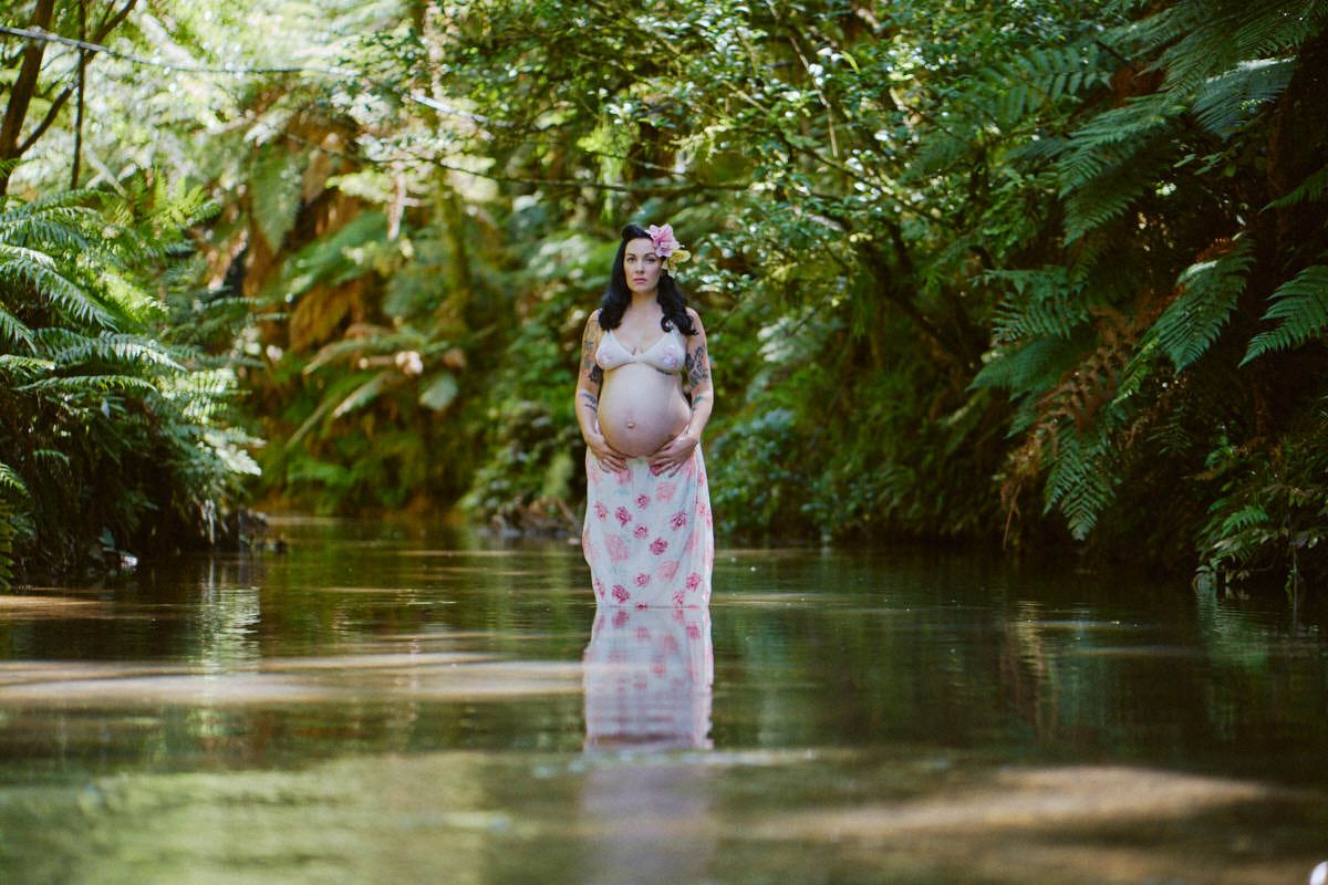 Pregnant woman in floral dress standing in a serene forest river surrounded by lush green tropical foliage.