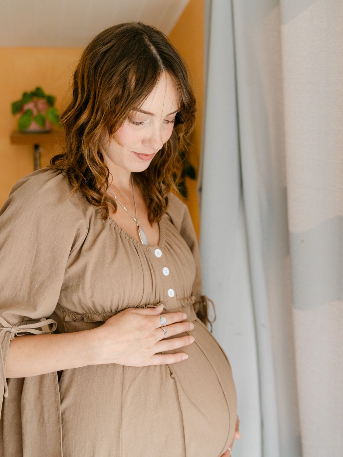 Pregnant woman in tan dress cradling her baby bump, standing by a window in soft natural light.