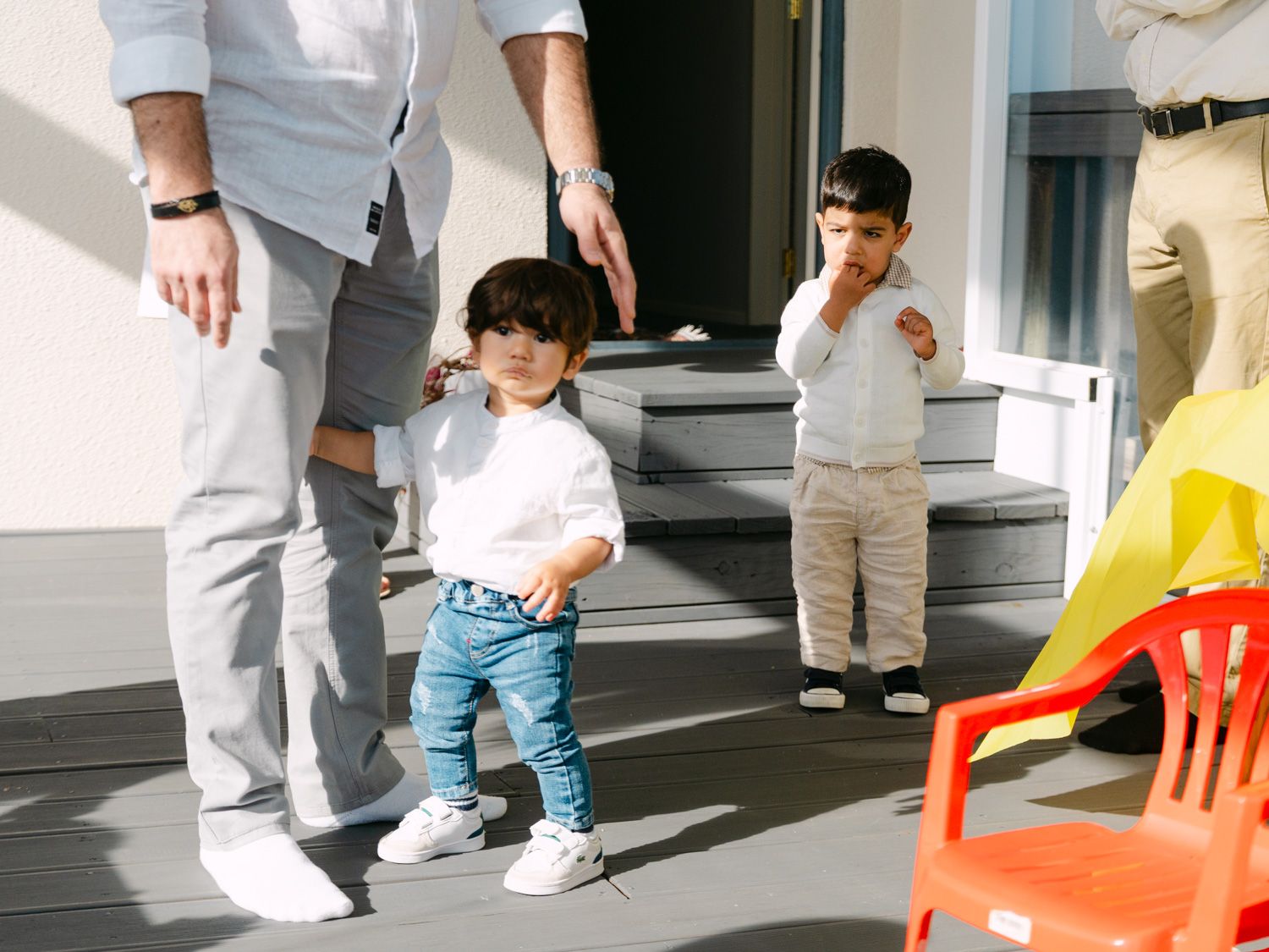 Two toddlers playing outdoors near house steps while an adult supervises on a sunny day.