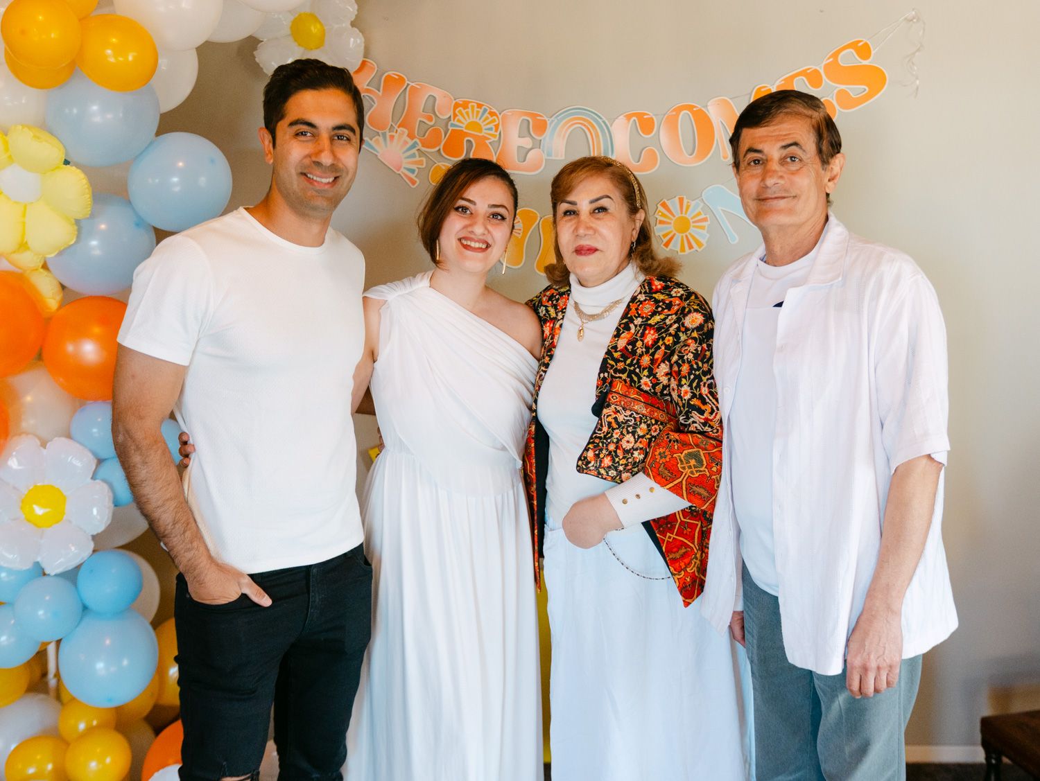 Four people posing together at a festive celebration with colorful balloon arch and party decorations in background.