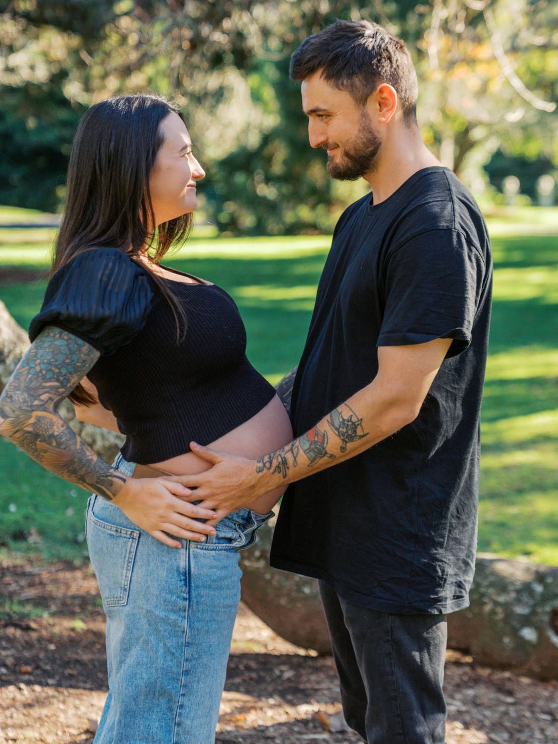 Pregnant couple in black outfits holding baby bump in sunny park, both tattooed, sharing a tender moment.
