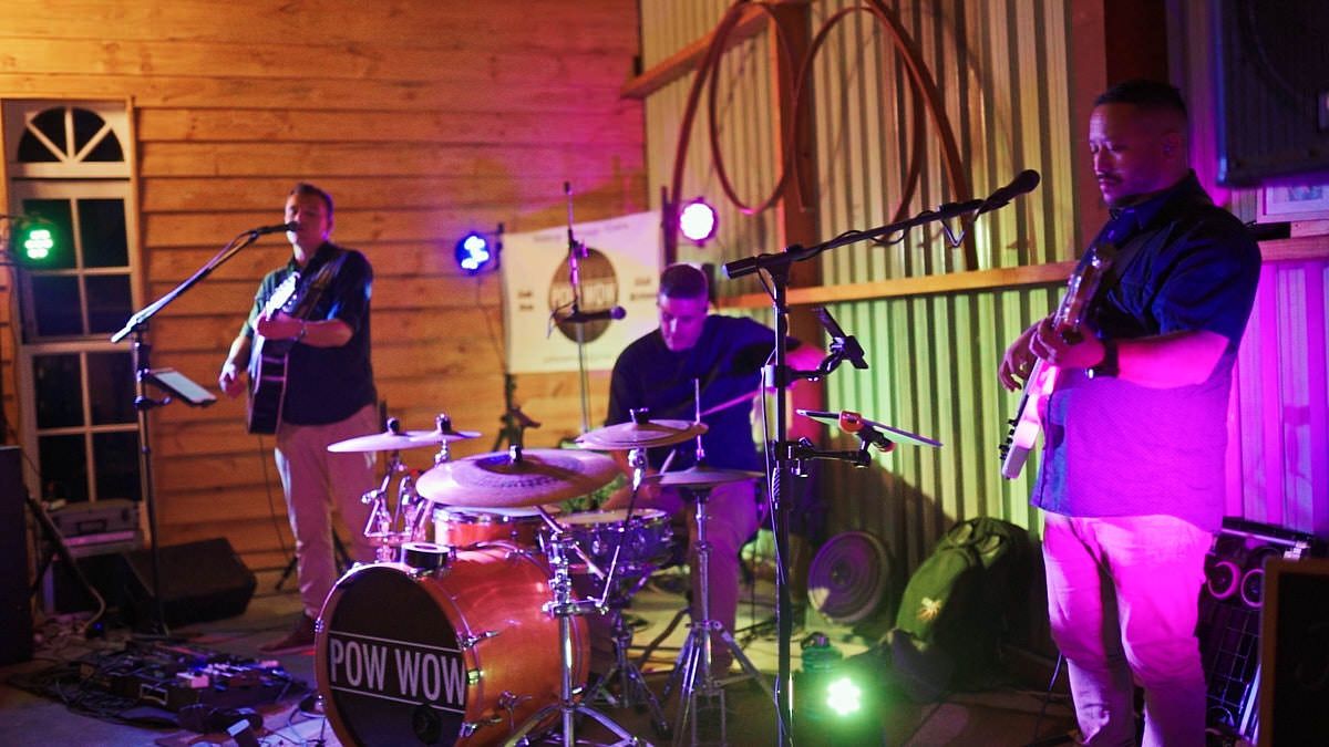 Live band performing on stage with drummer and guitarist in a rustic venue with purple and pink lighting.