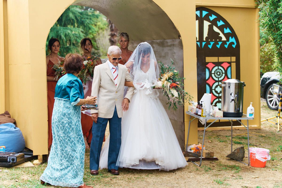 Bride in white gown with veil walks through yellow arch with elderly escort at outdoor wedding ceremony.