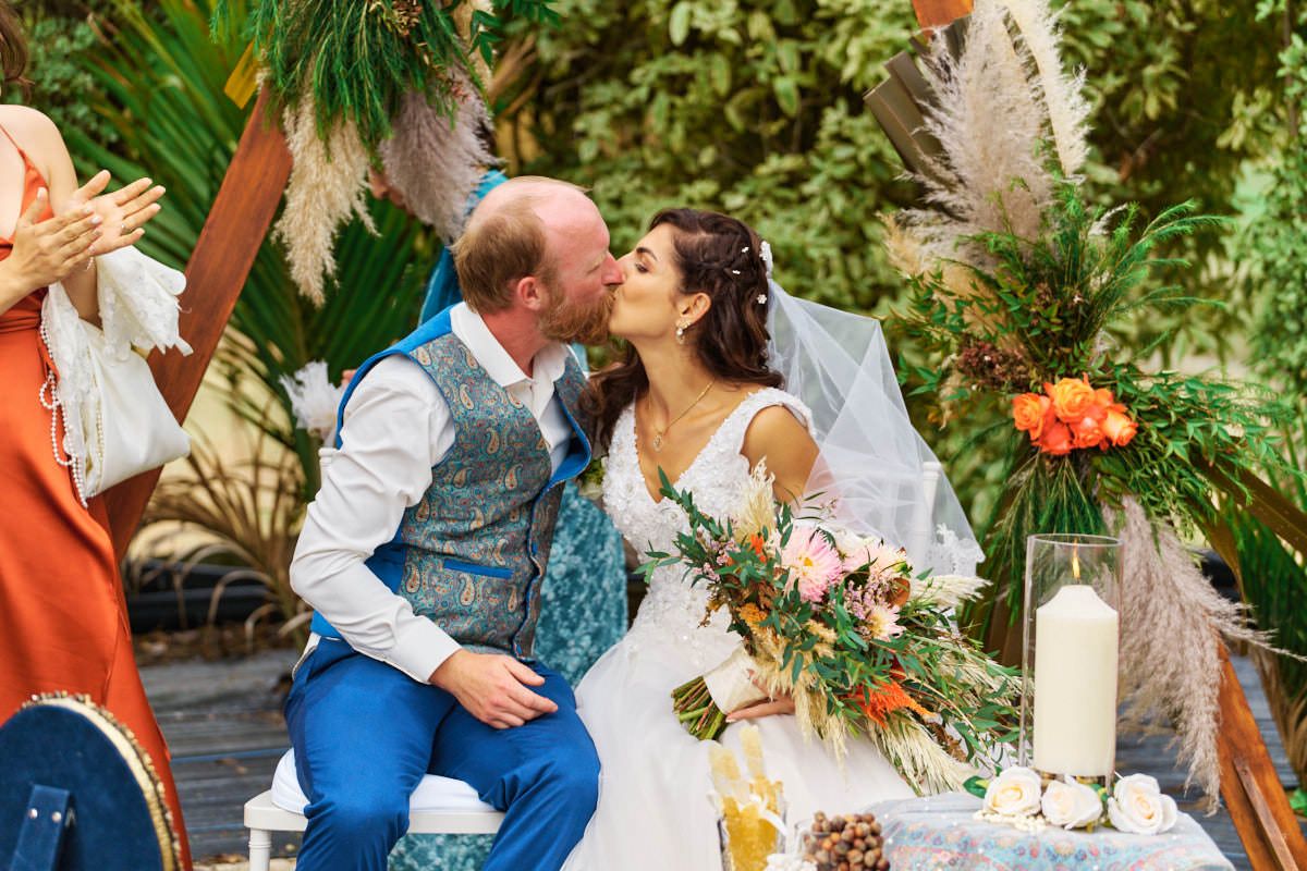 A bride and groom share a romantic kiss at their boho outdoor wedding ceremony with tropical greenery and colorful floral decor.