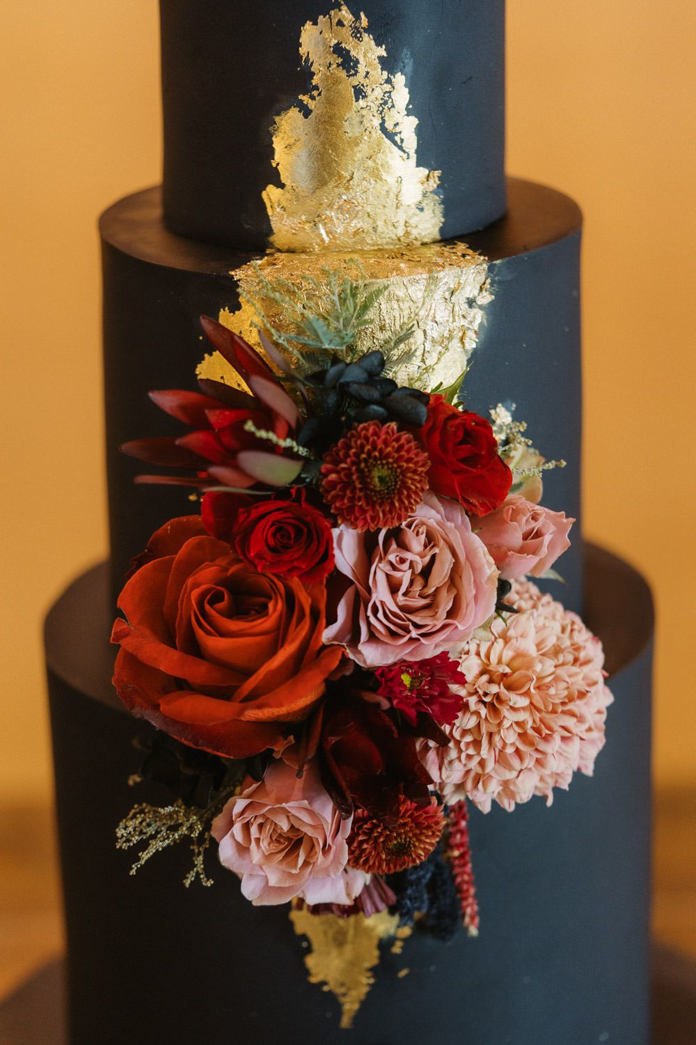 Elegant three-tier black wedding cake adorned with gold leaf and vibrant red, pink, and burgundy floral arrangement.