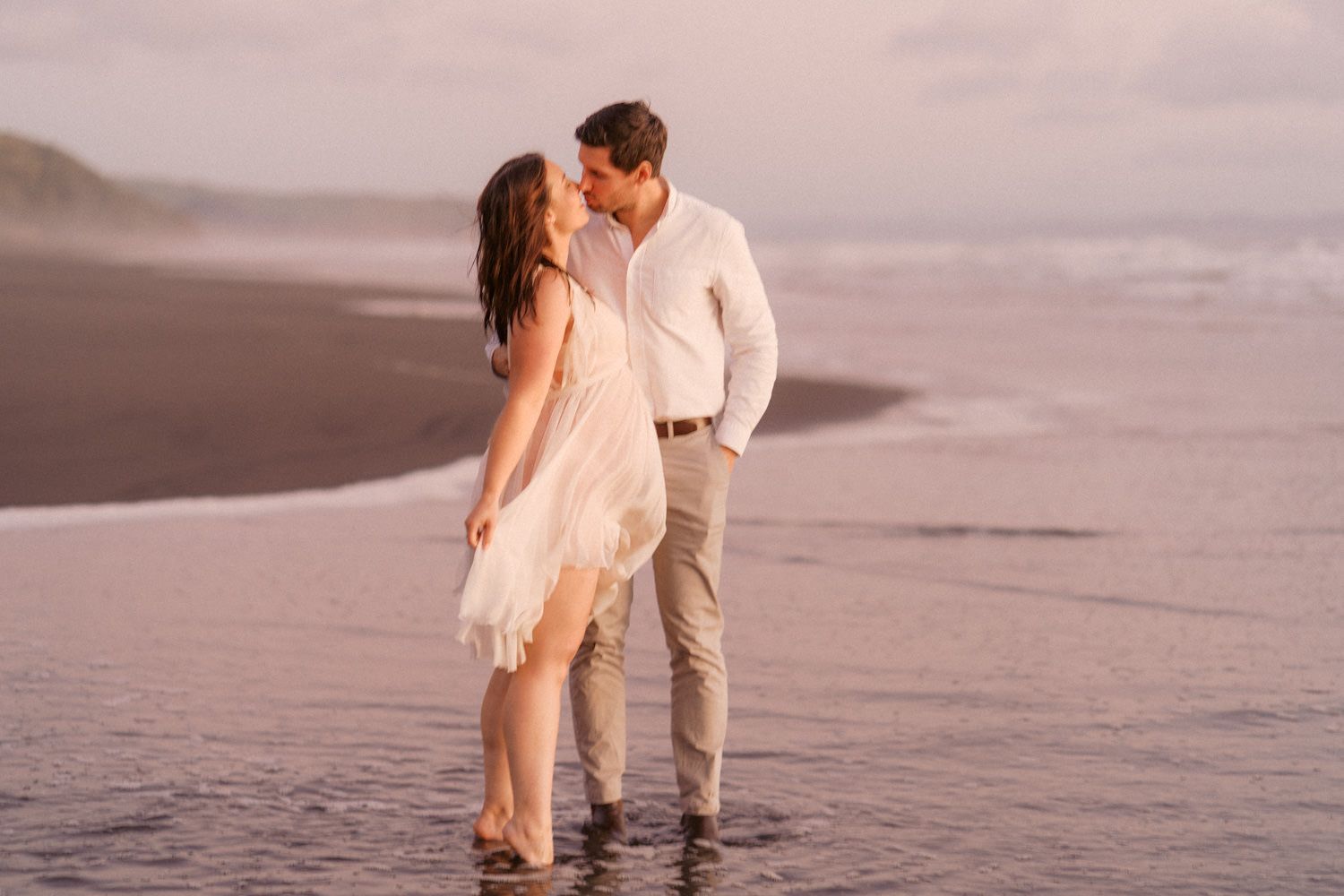 Couple kissing on a sandy beach at golden sunset, woman in white dress, man in white shirt and khaki pants.