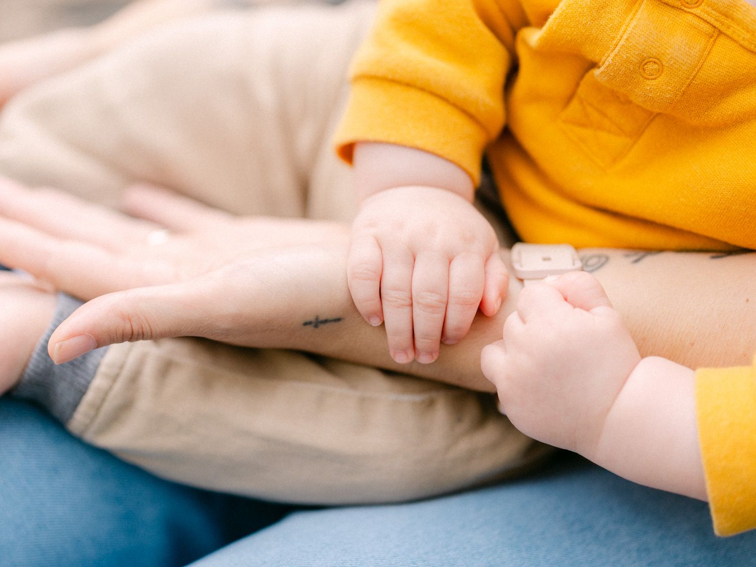 Close-up of a baby in yellow outfit gently holding an adult's hand with a small arrow tattoo on the wrist.