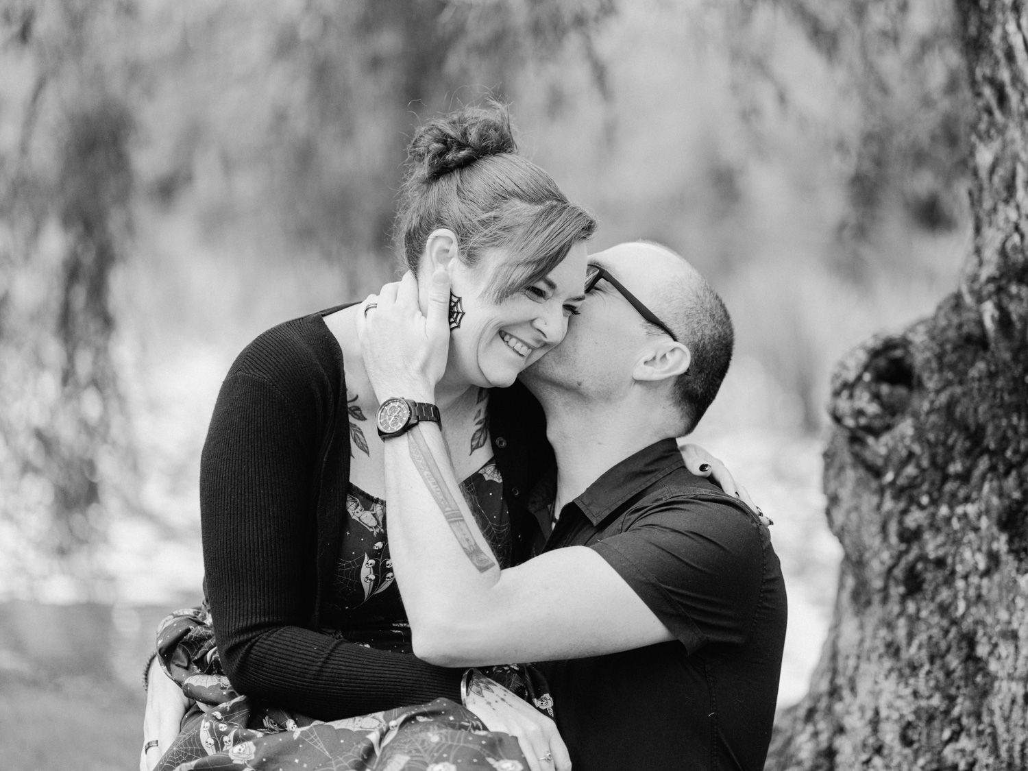 Black and white photo of a couple sharing a romantic moment outdoors, man kissing womans cheek as she smiles.