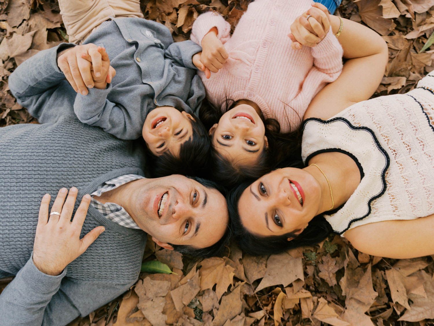 Four family members lying on autumn leaves smiling up at camera in a joyful outdoor fall portrait.