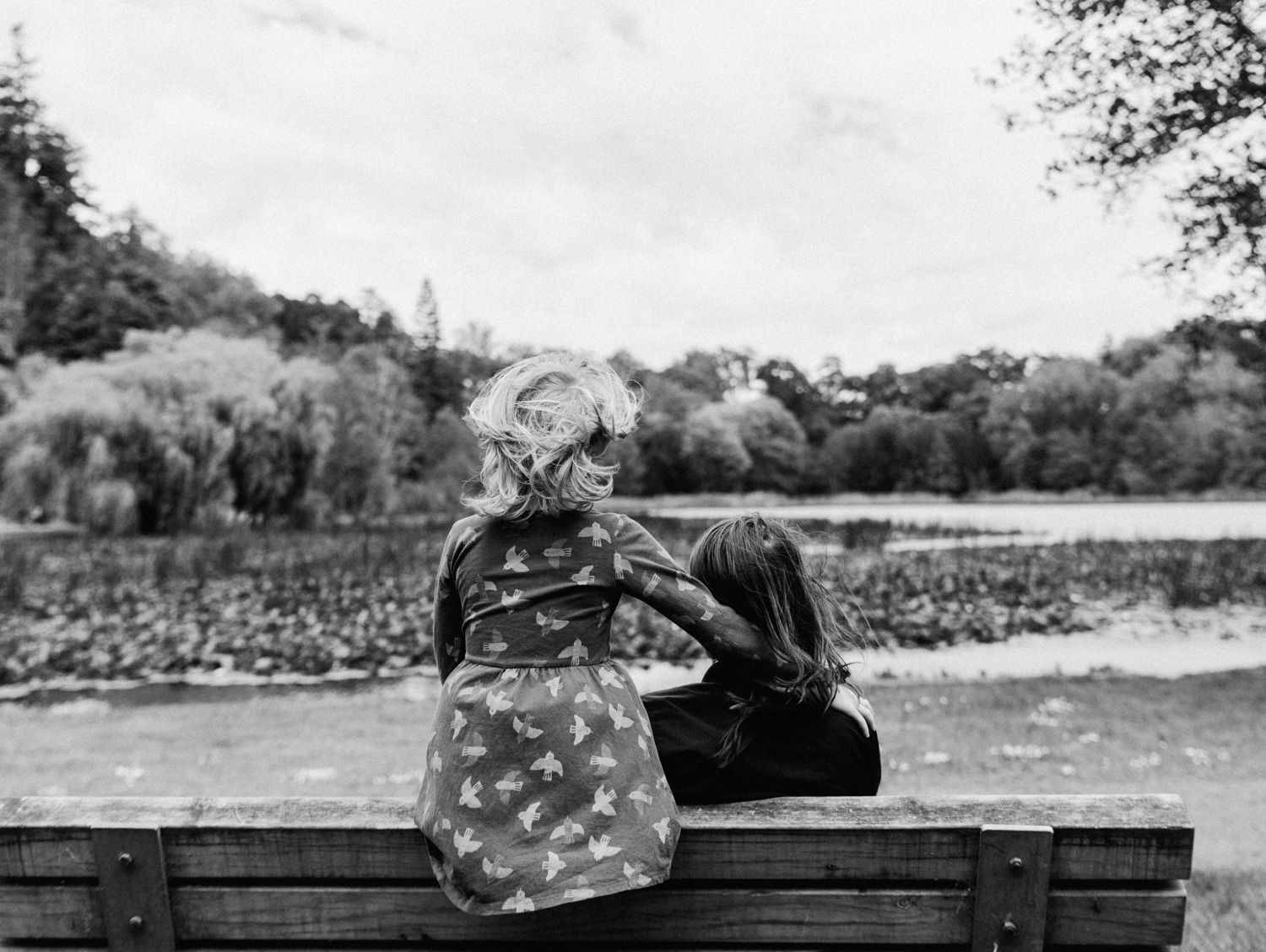 Two children sit together on a wooden bench, sharing a quiet moment by a scenic park lake in black and white.