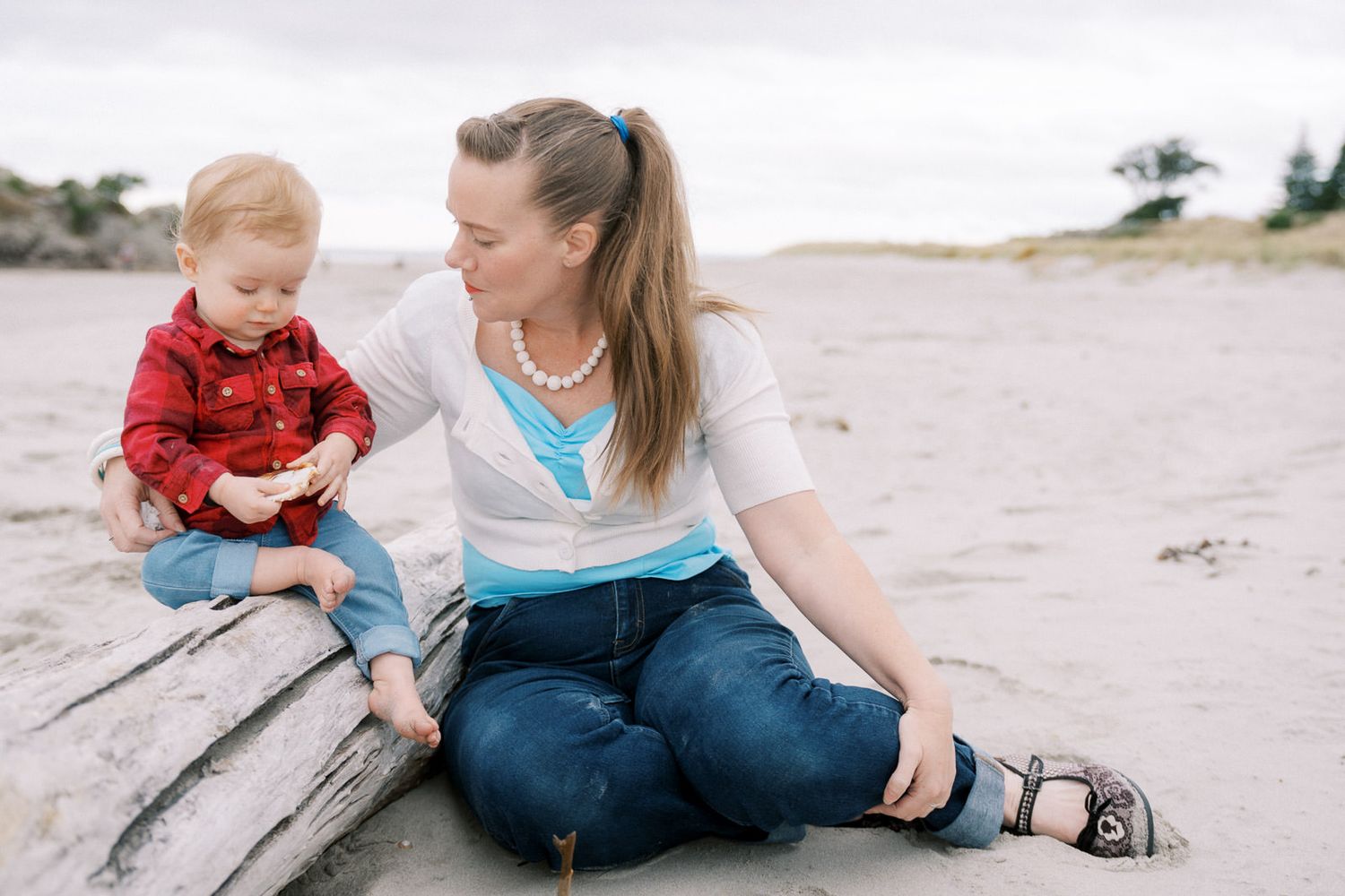 A mother and toddler sit on driftwood at a beach, sharing a tender moment while the child holds a small object.