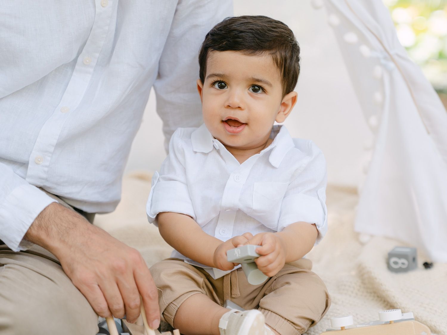 A cute toddler boy in a white shirt and khaki pants sits on a blanket near a teepee, playing with toys.