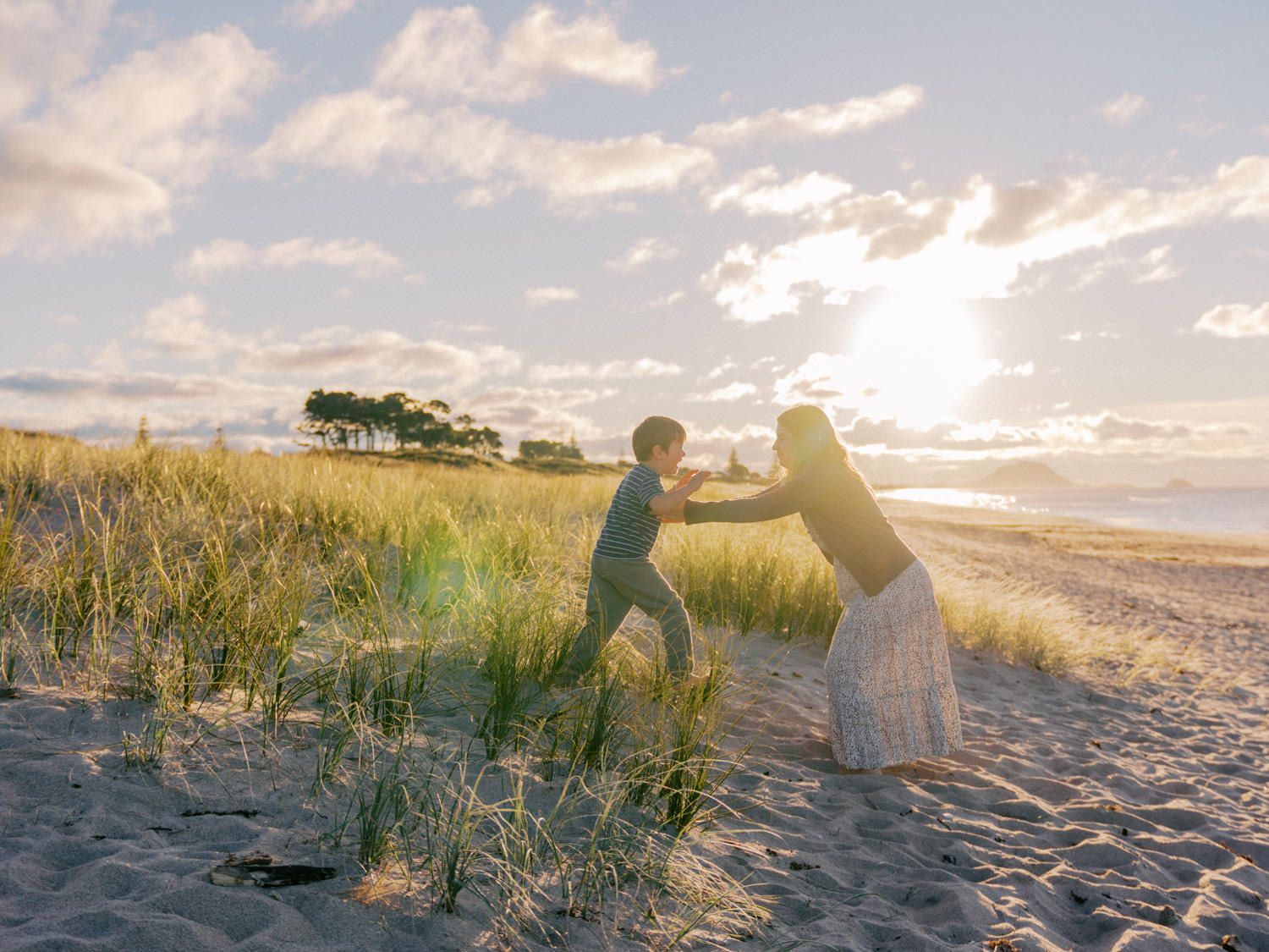 Two people playfully reaching toward each other on a sandy beach at golden sunset with grassy dunes in the background.