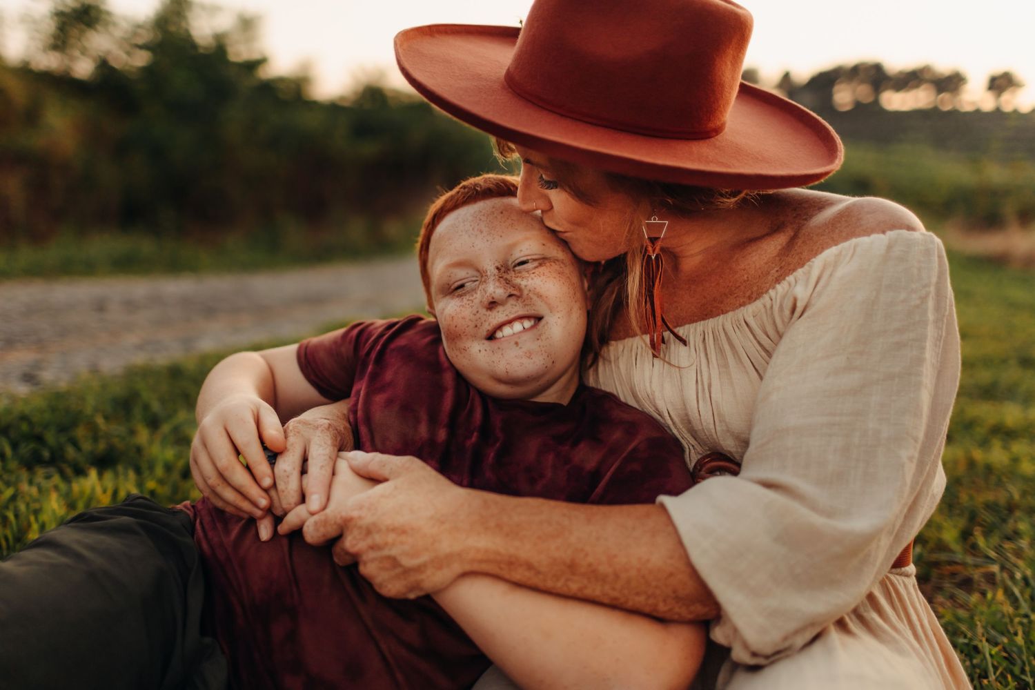 A woman wearing a rust hat tenderly kisses a laughing red-haired boy while hugging him on grass.