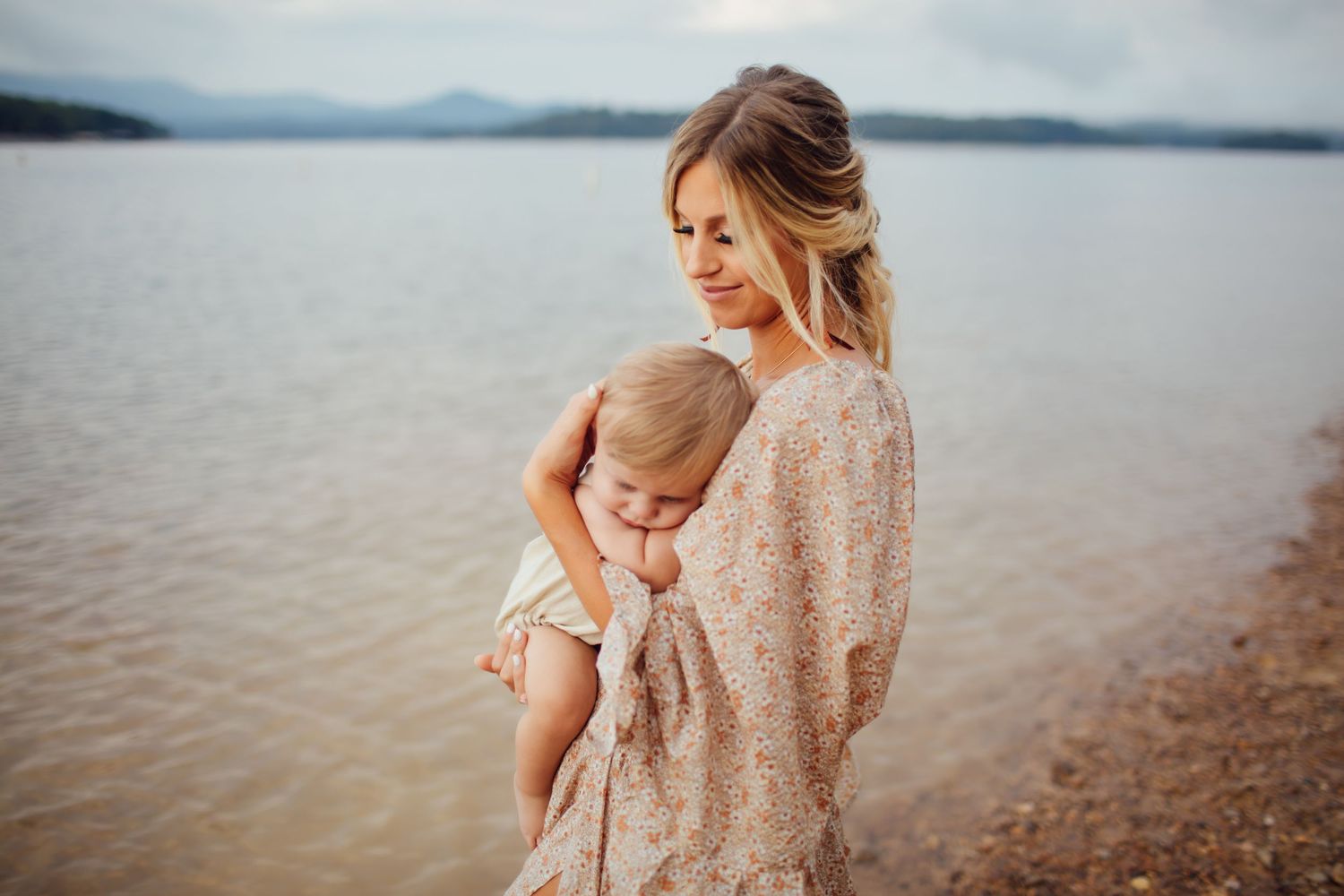 A young mother in a floral dress tenderly holds her sleeping baby by a calm lake.