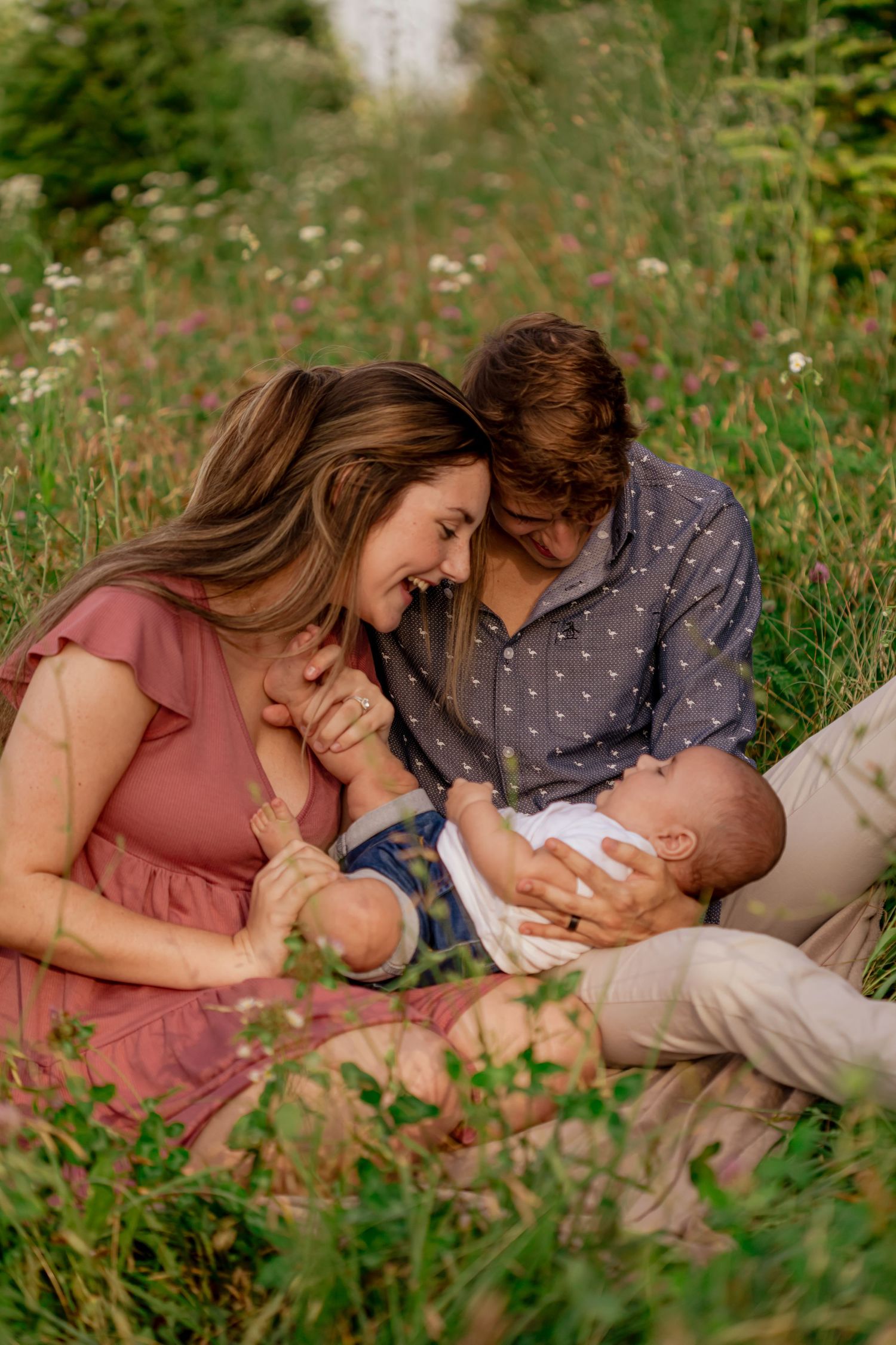 A couple sits in a wildflower field, smiling at each other while cradling their newborn baby outdoors.