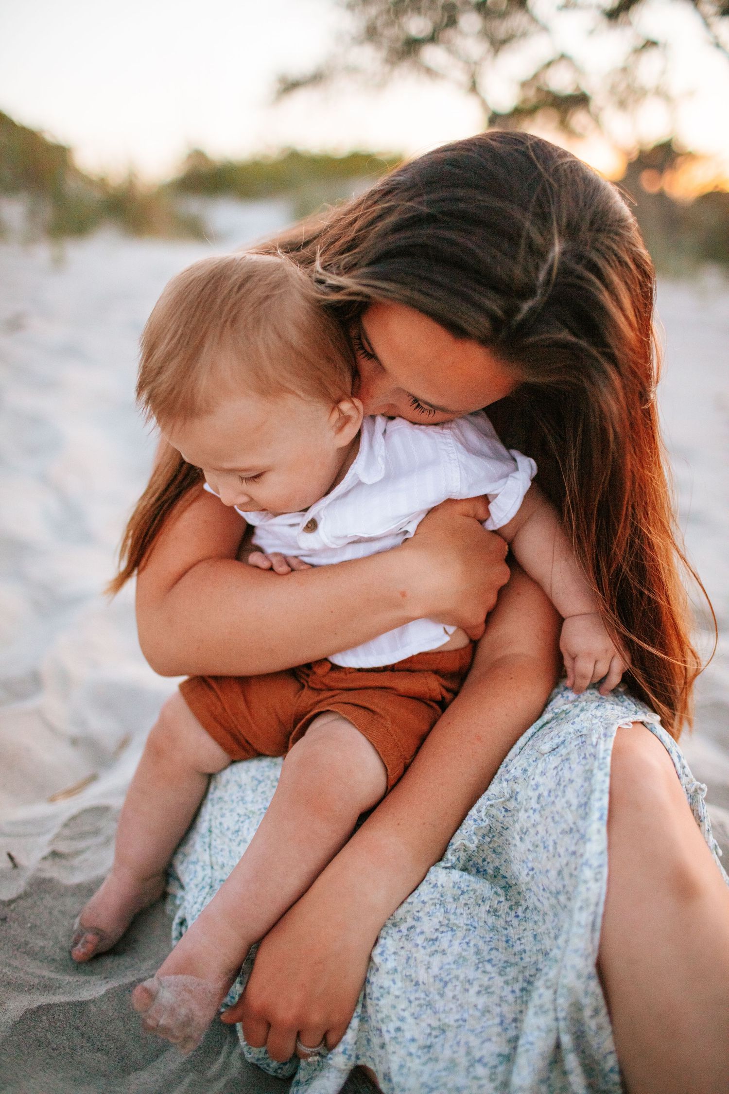Mother tenderly hugging her baby on a sandy beach at golden hour sunset.
