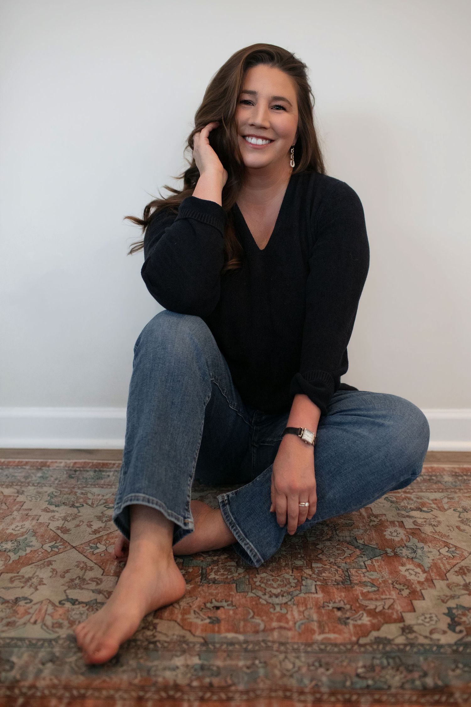Woman in black top and blue jeans sitting barefoot on a vintage rug, smiling and touching her hair.