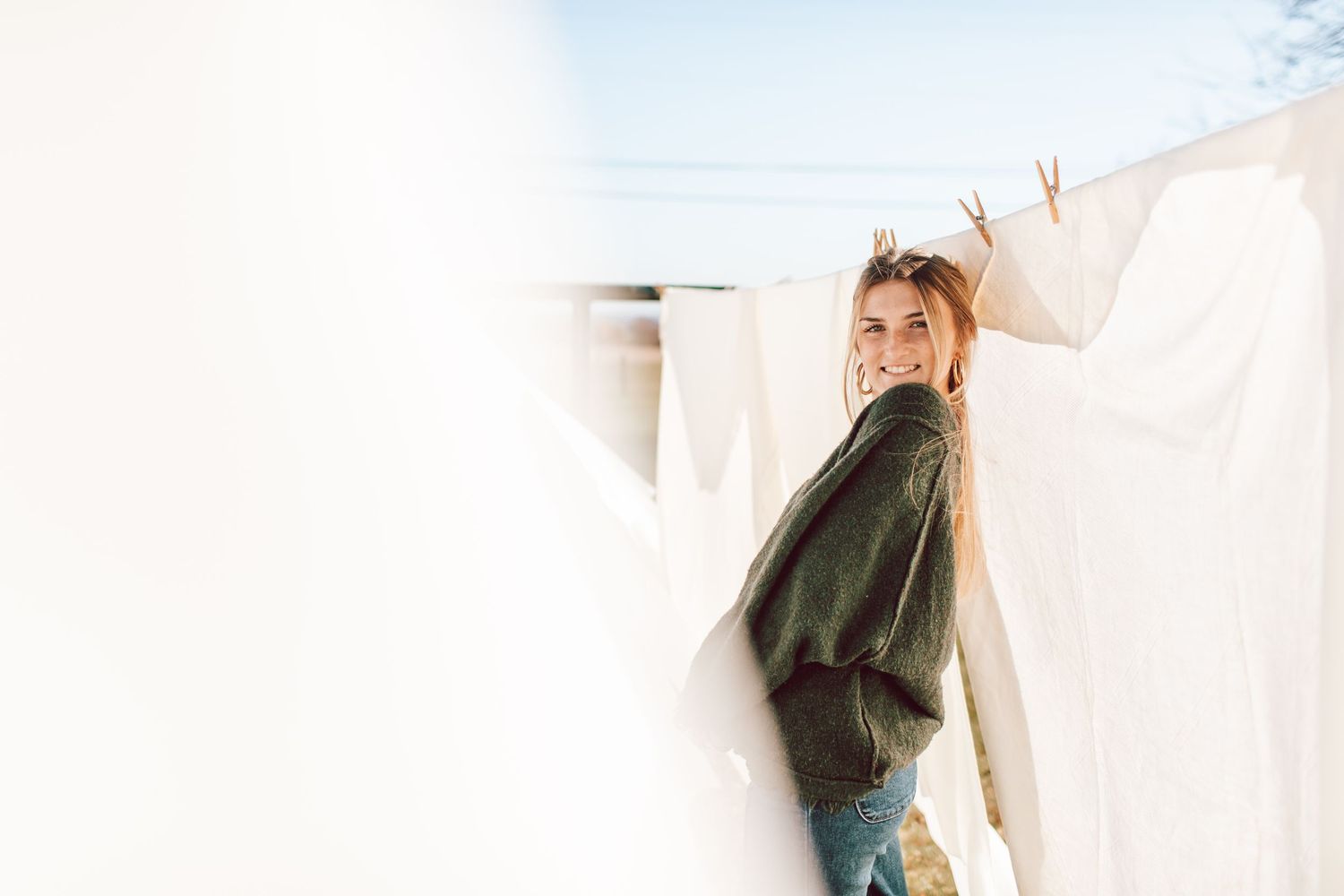 Smiling blonde woman standing between white sheets hanging on a clothesline outdoors on a sunny day.
