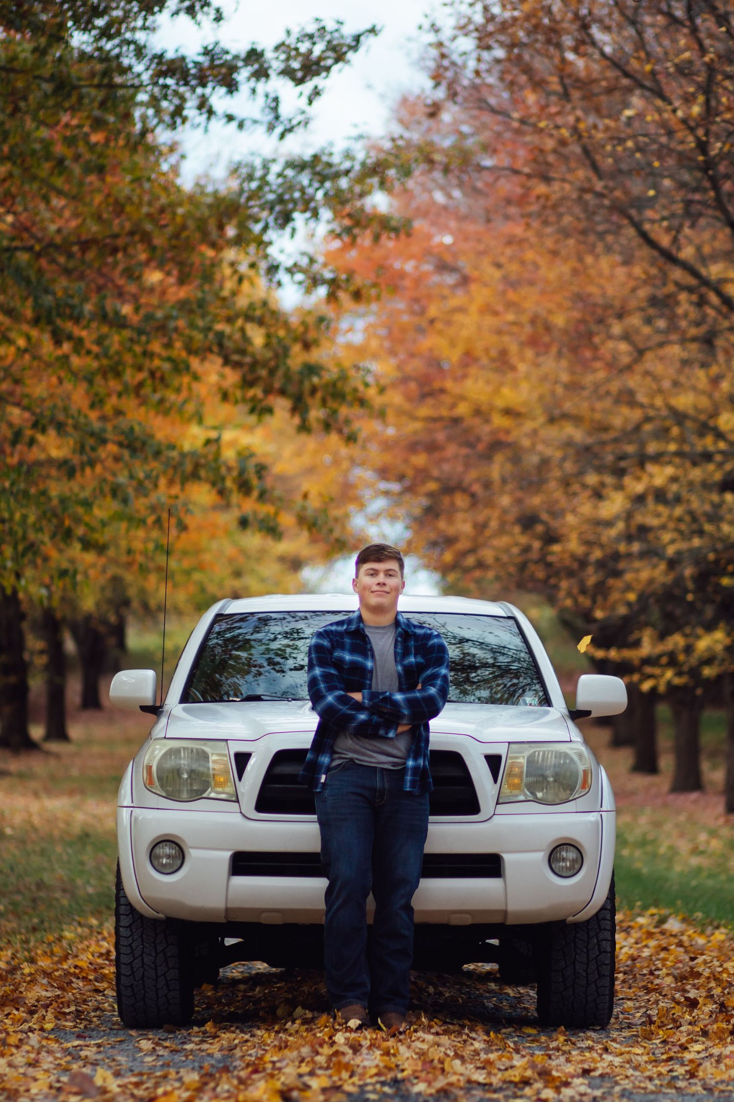 A young man in a blue plaid shirt poses in front of a white Toyota Tacoma on a fall foliage road.