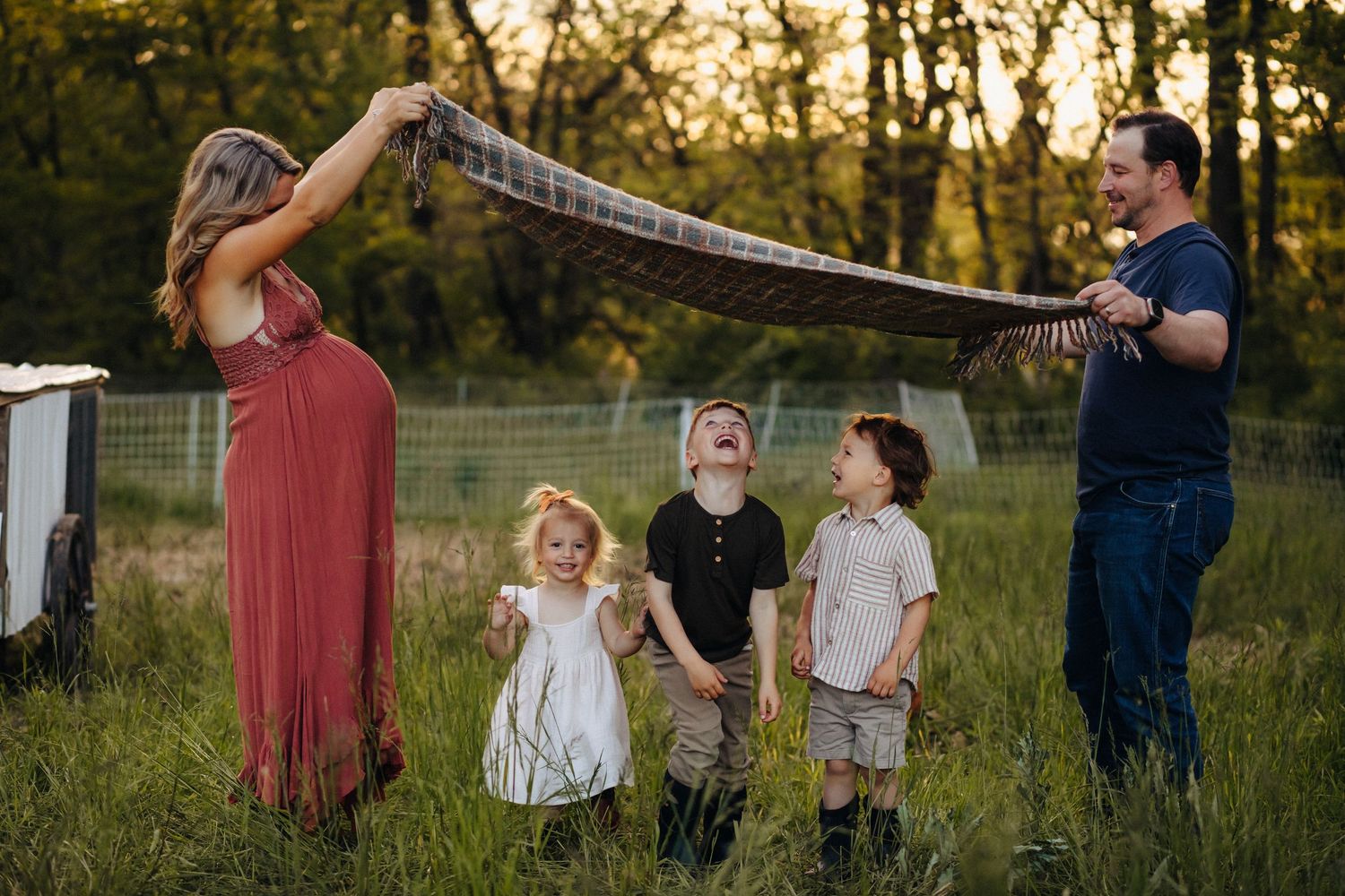 Family of five playing outdoors in a meadow at sunset, parents lifting a blanket over laughing children.