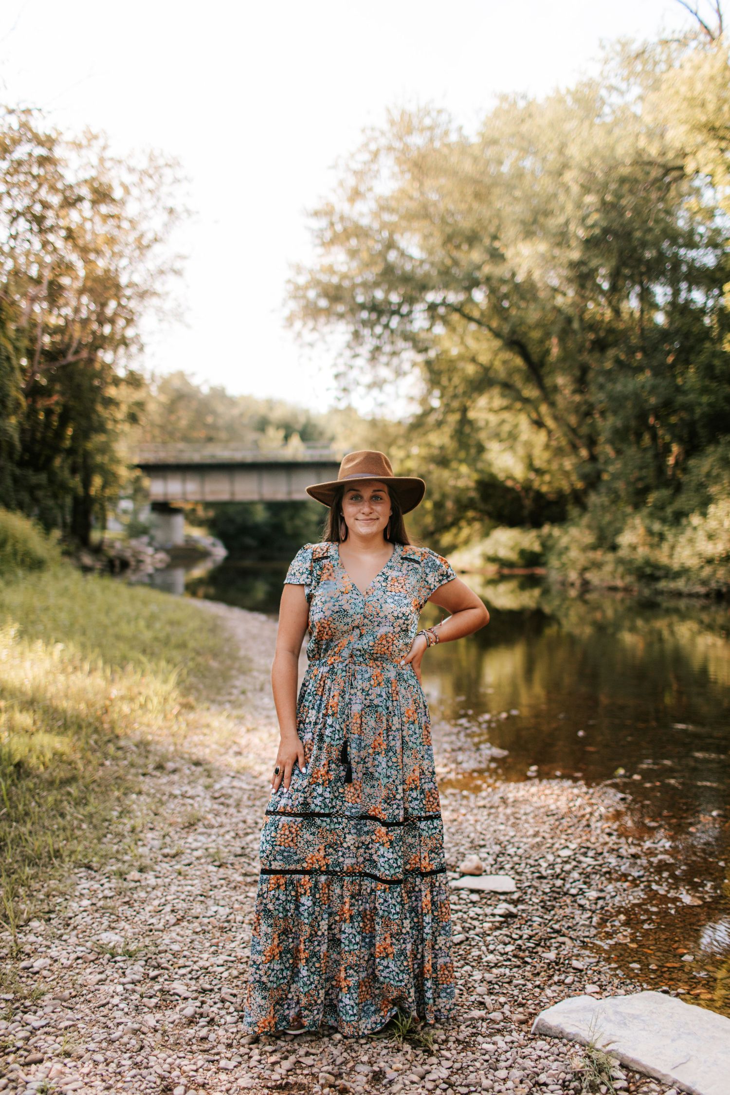 Woman in floral maxi dress and hat standing by a river near a covered bridge.