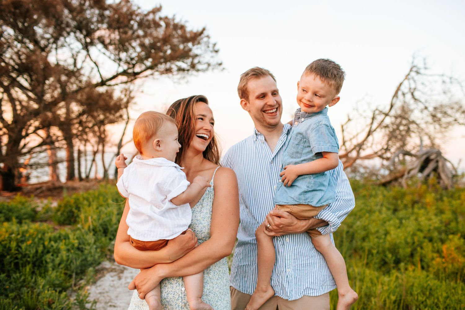 A joyful family of four smiles together outdoors at sunset, with parents each holding a young child near trees.