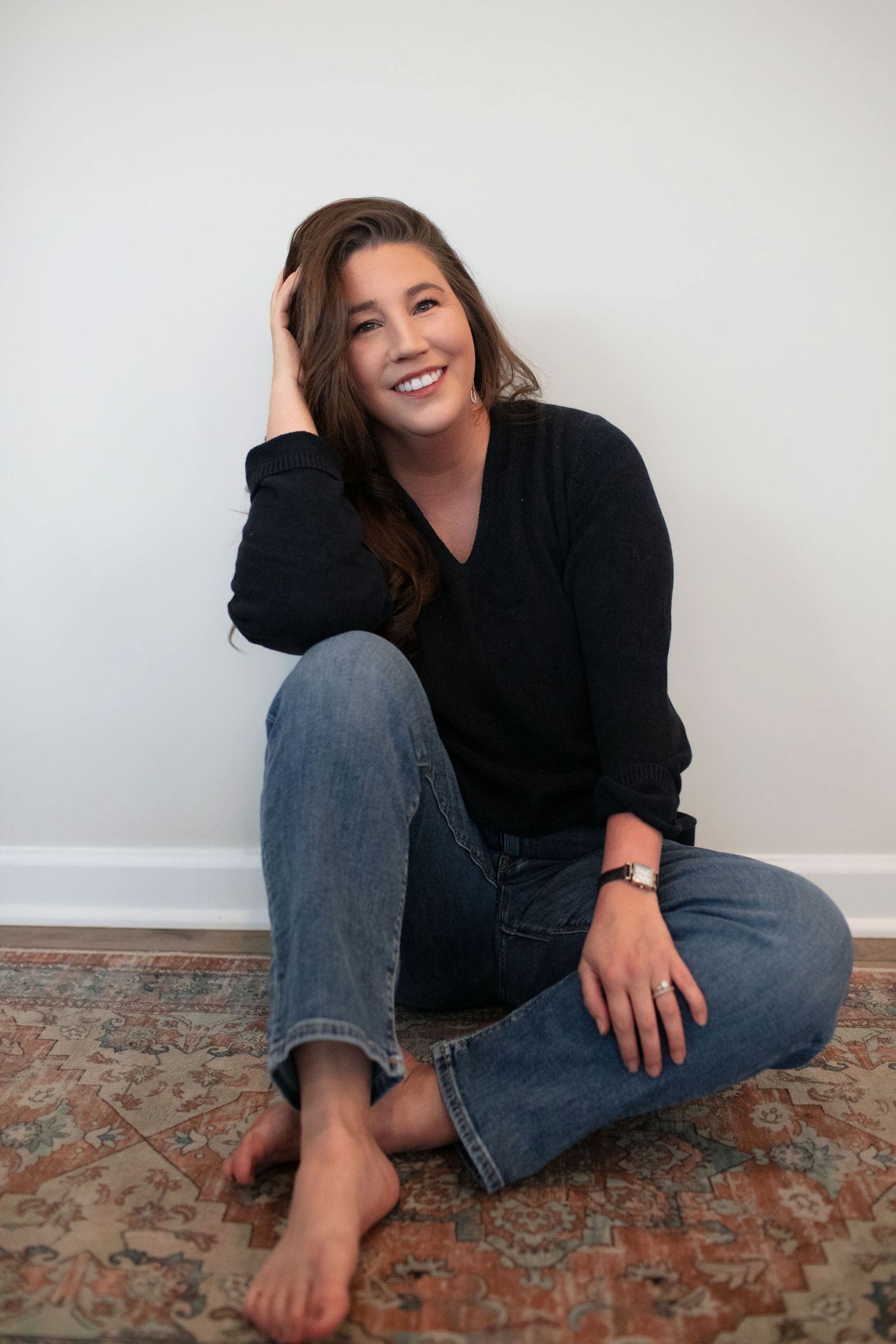 Brunette woman in black sweater and jeans sitting on a rug, smiling and resting hand on head.
