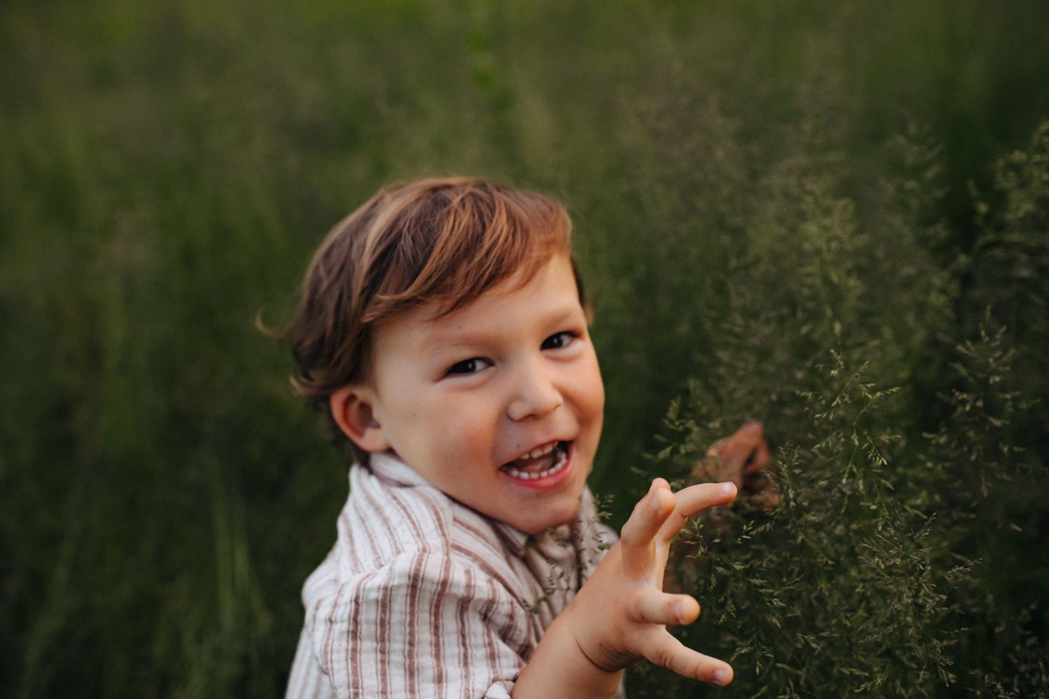 Happy toddler boy smiling in a lush green field, wearing a striped shirt while reaching out to touch plants.