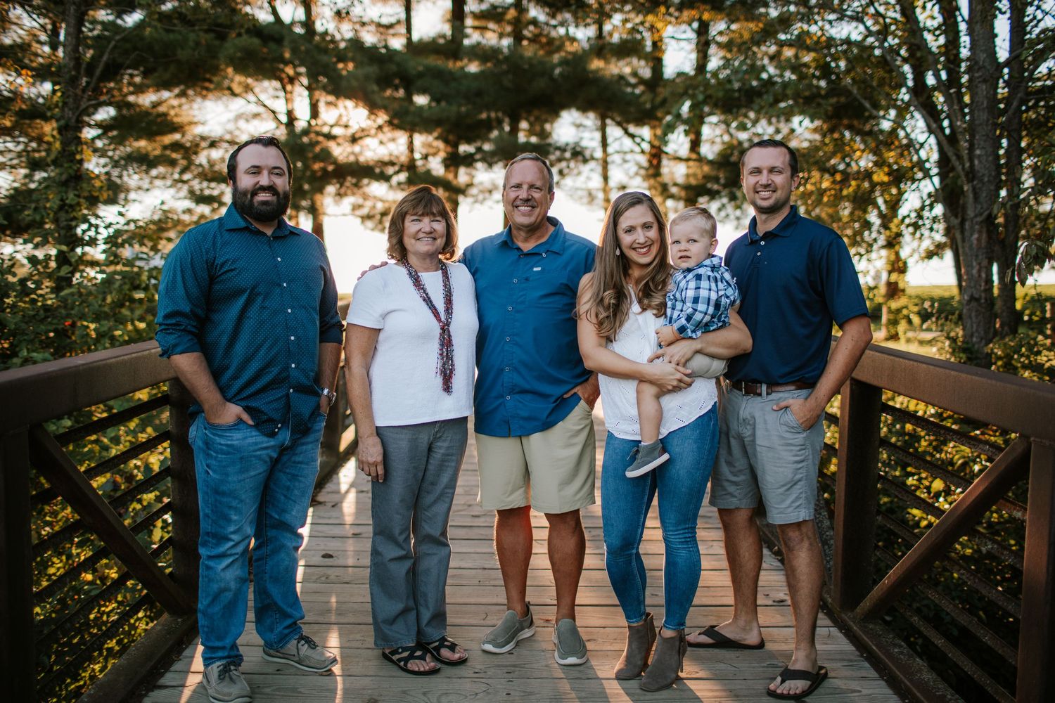 Extended family of six smiling together on a wooden bridge surrounded by trees.