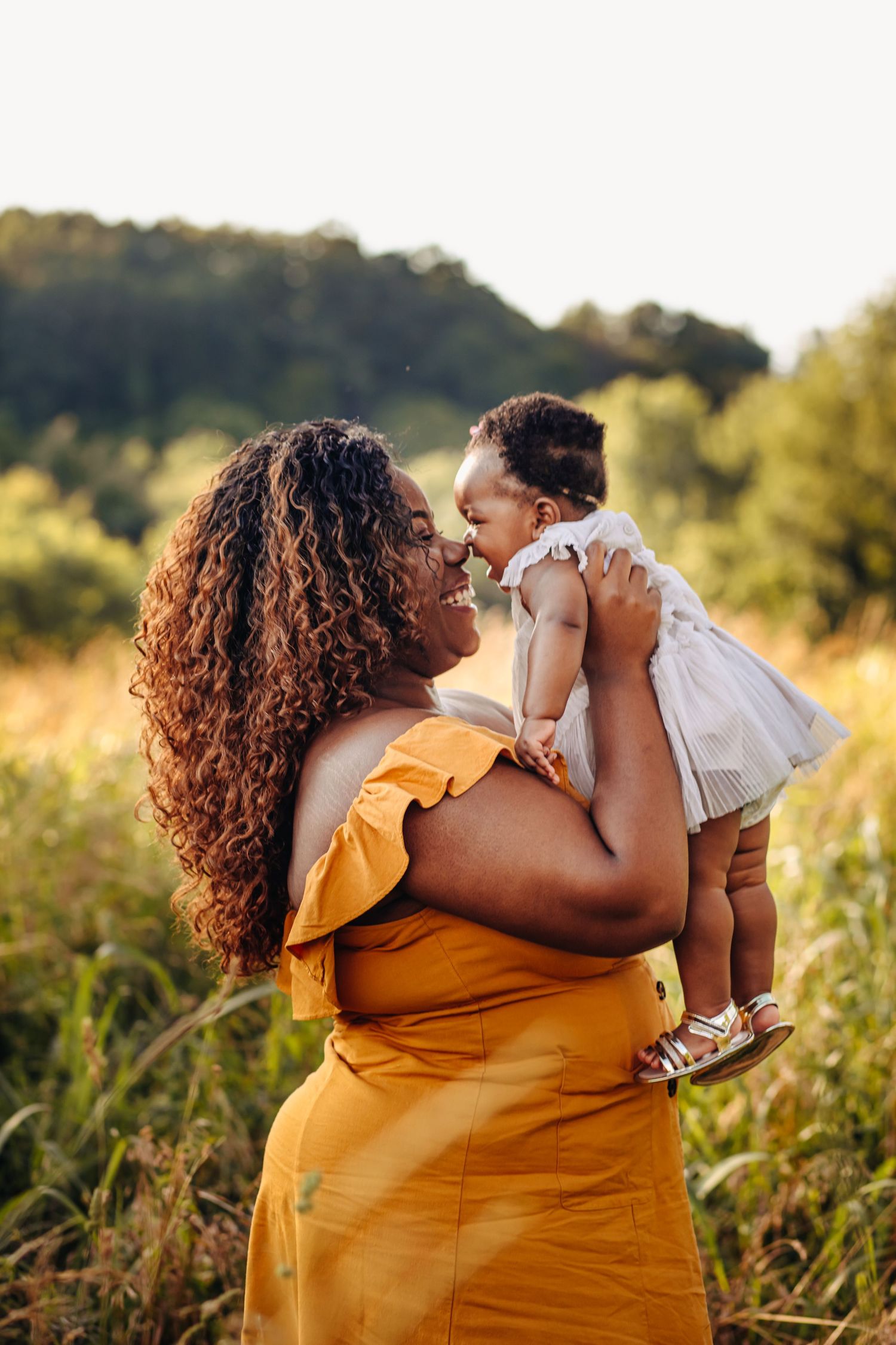 A joyful mother in a yellow dress holds her baby girl up in a sunny green field.