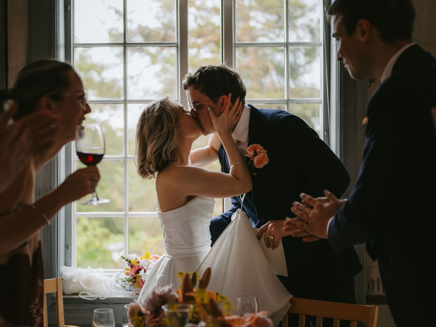 Bride and groom sharing kiss at wedding reception table with guests.