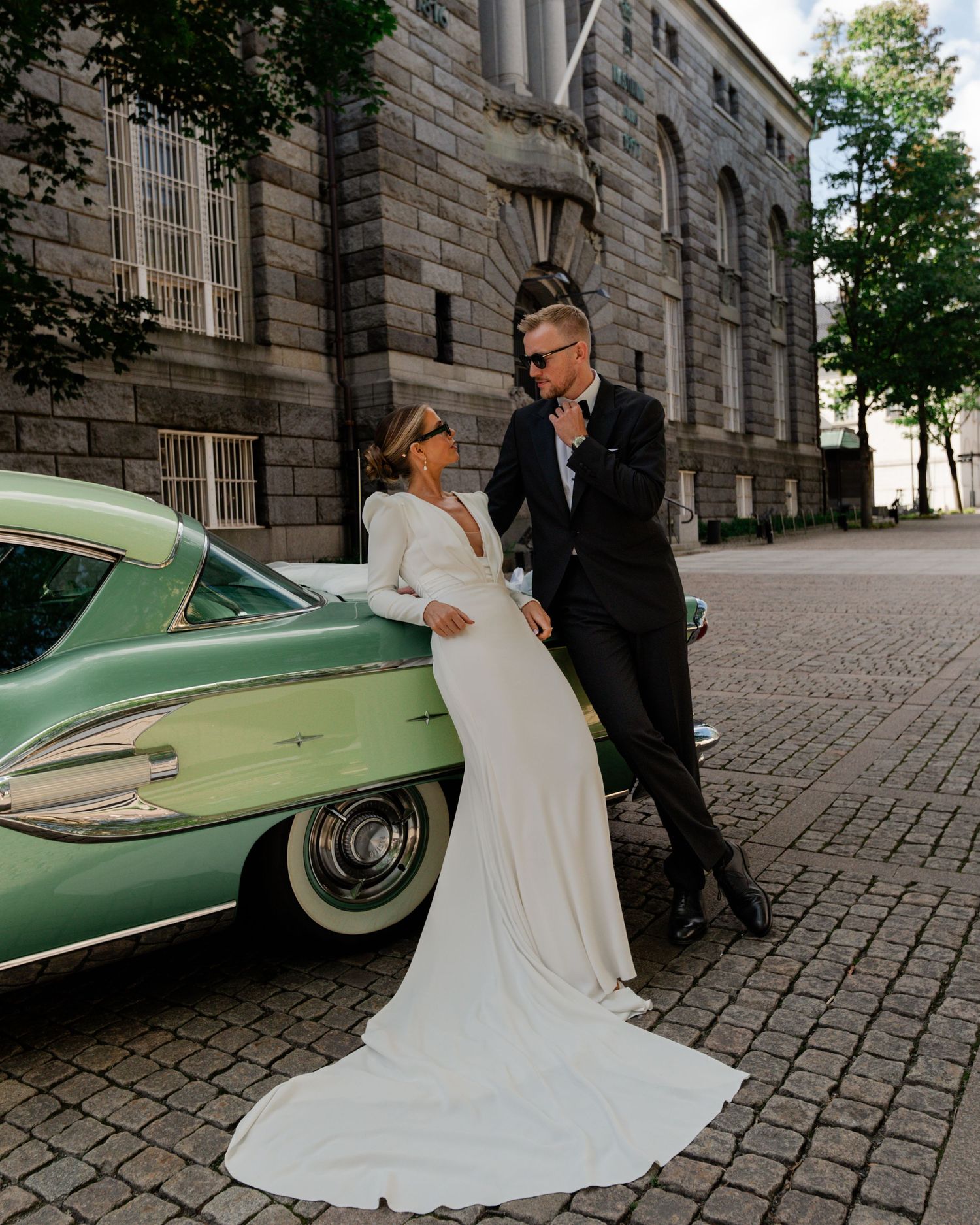 Chic bride and groom posing beside vintage green car outside building.