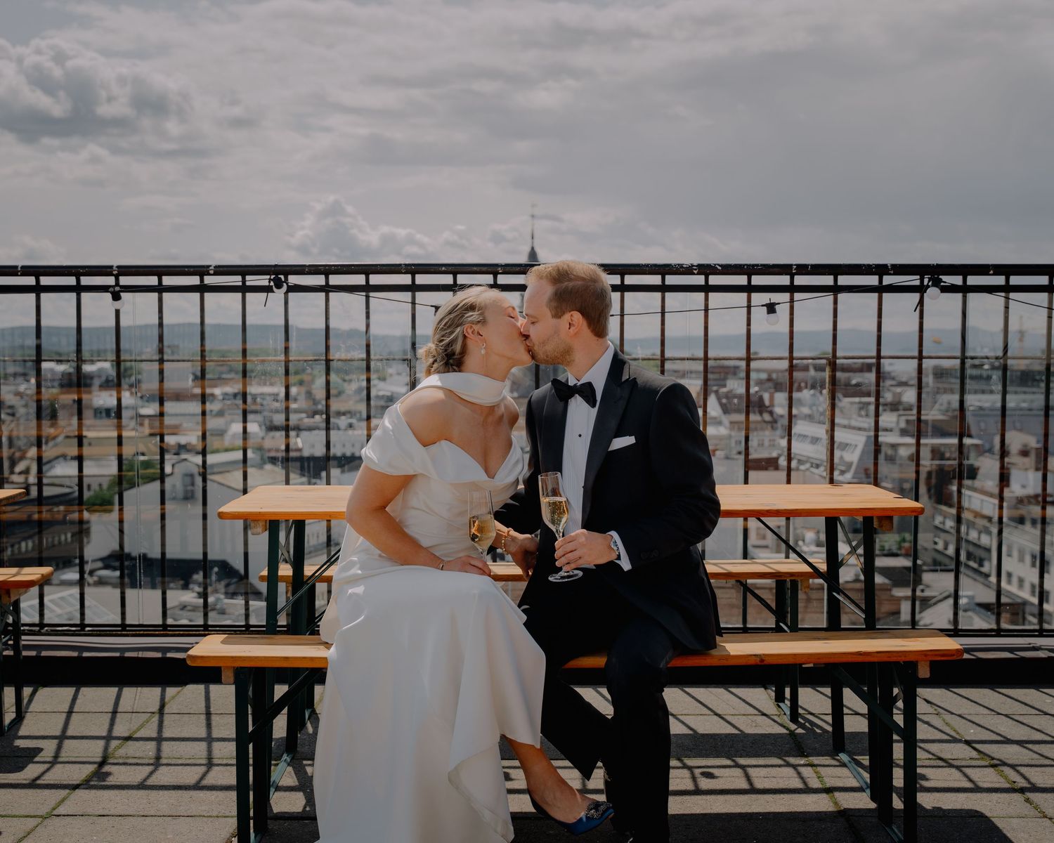 Newlyweds kissing on rooftop terrace with champagne and city panorama.