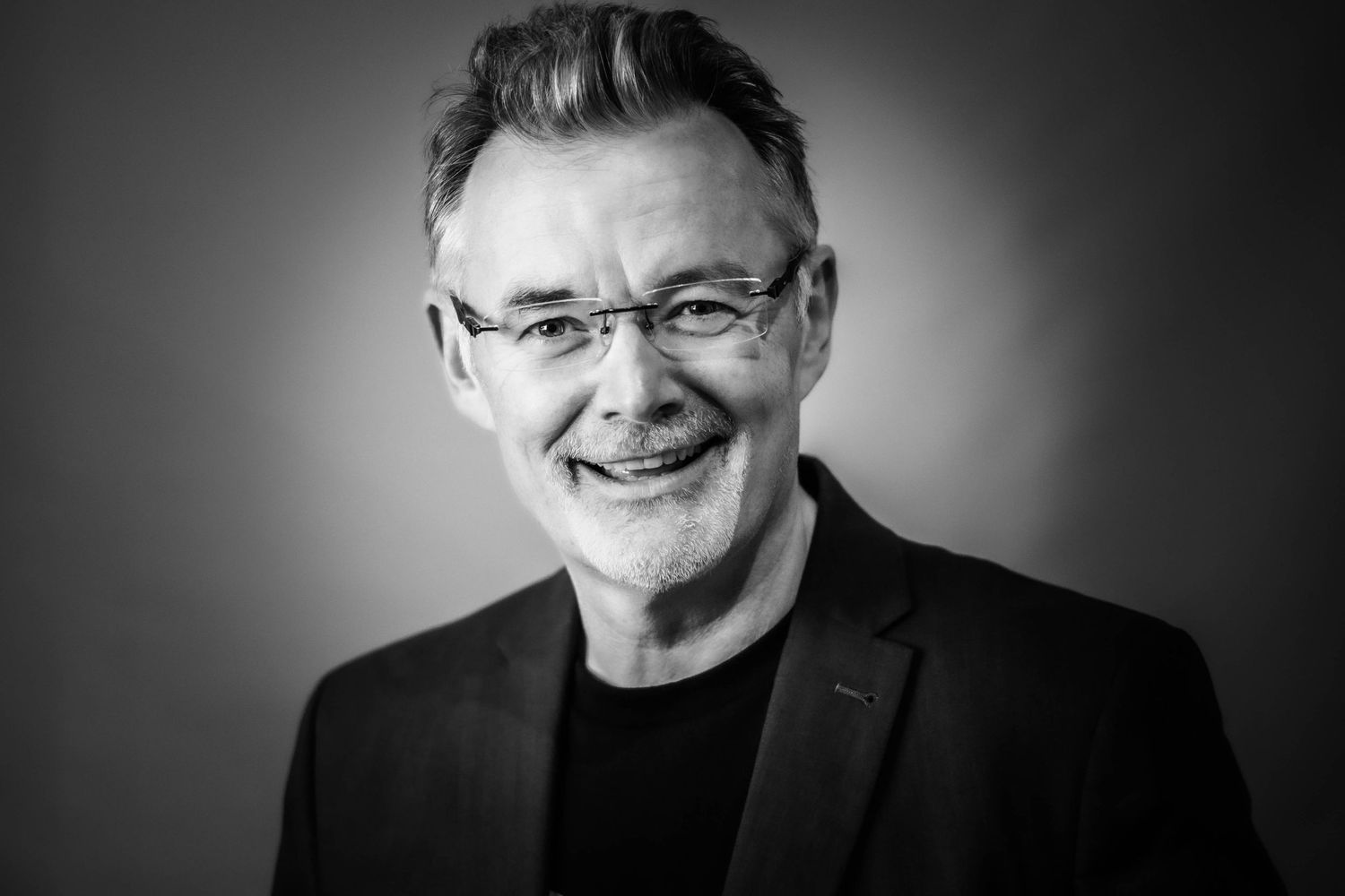 Professional studio black and white headshot of a smiling man in a navy blue suit against a solid grey background.