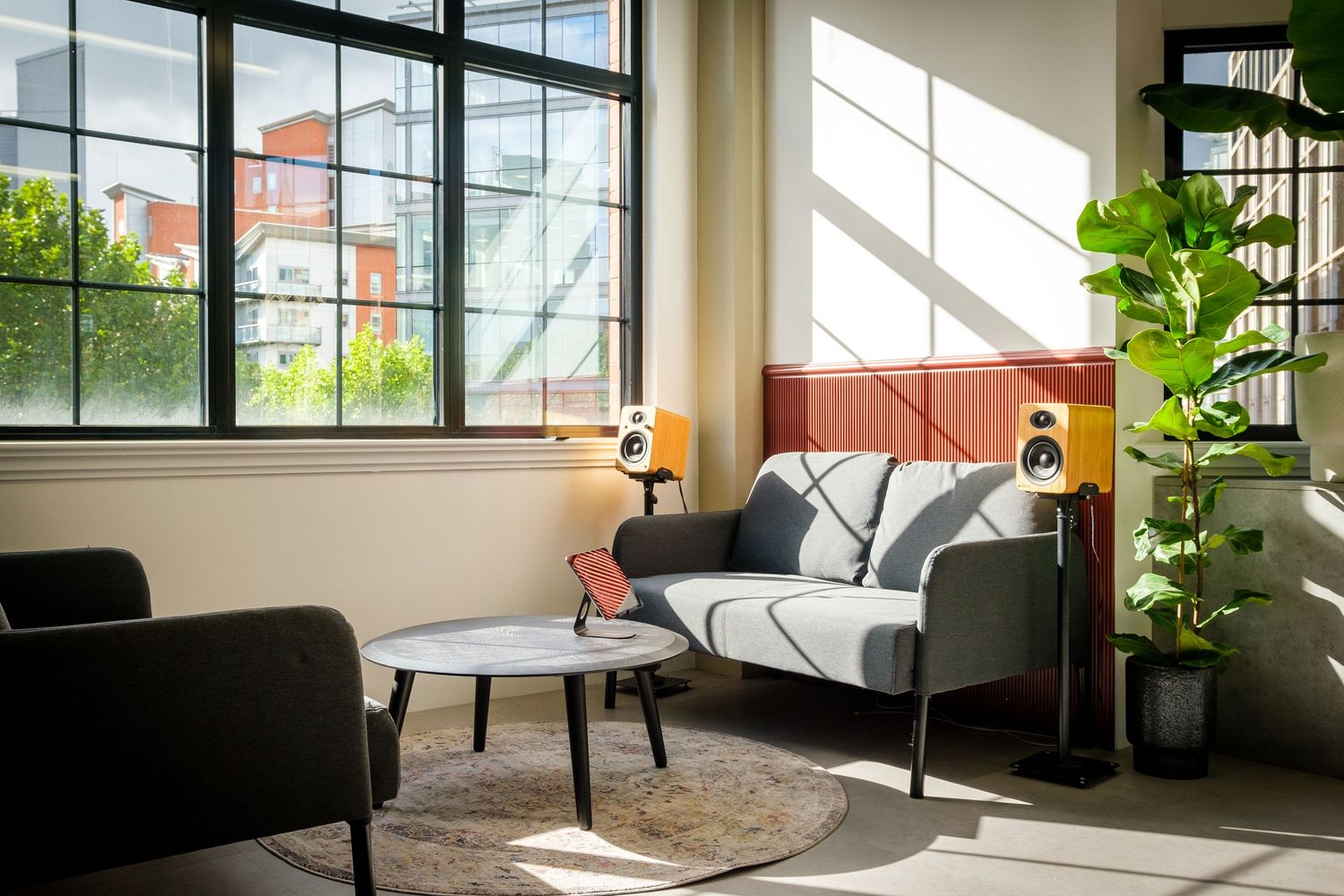 Sunlit office lounge with a grey sofa, round coffee table, speakers and a tall leafy plant, by Simon Robertson.
