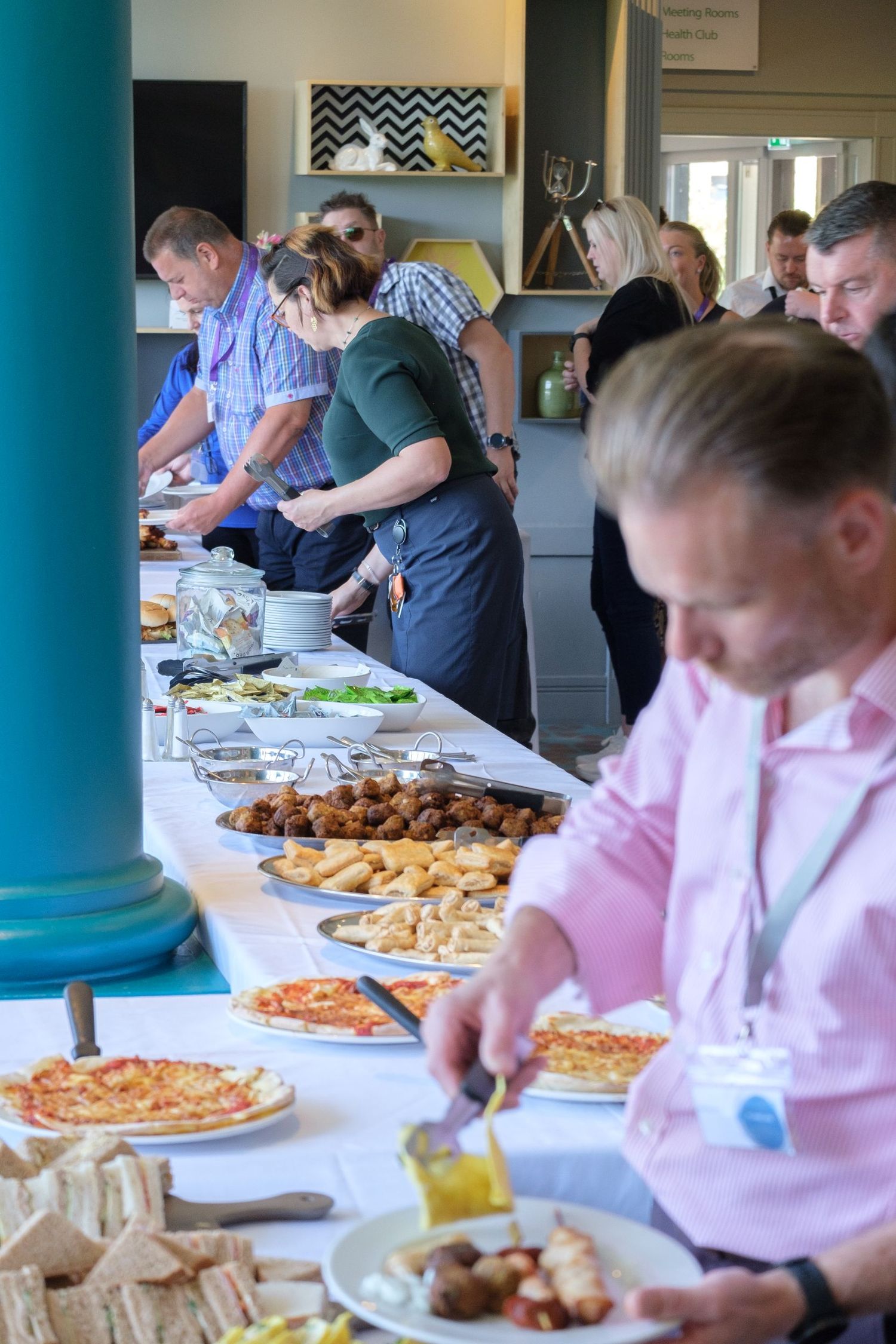 Guests helping themselves to a buffet of sandwiches, pizza and hot food at a reception, by Simon Robertson.