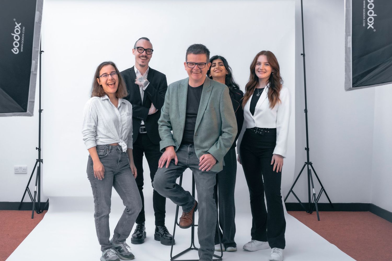 Five smiling professionals pose playfully in a studio, capturing a fun, relaxed, and modern corporate team culture.