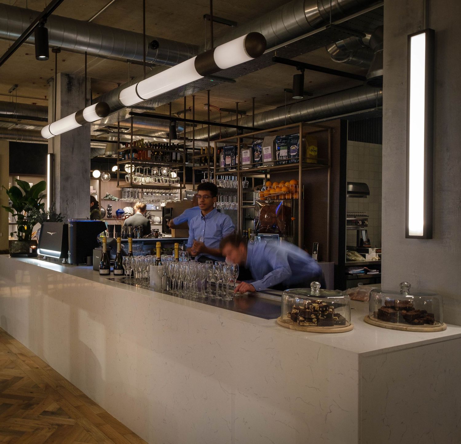 Bar staff behind a white counter beneath industrial pendant lights and exposed ductwork, by Simon Robertson.