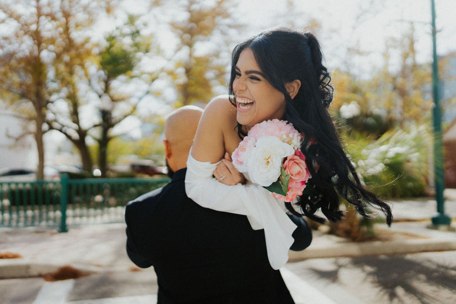 A newly married couple shares a romantic embrace outdoors while holding a pink and white bouquet.