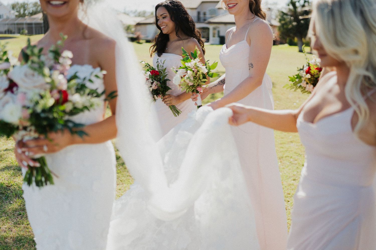 A wedding party in white dresses stands on grass outdoors holding vibrant floral bouquets in the sunlight.