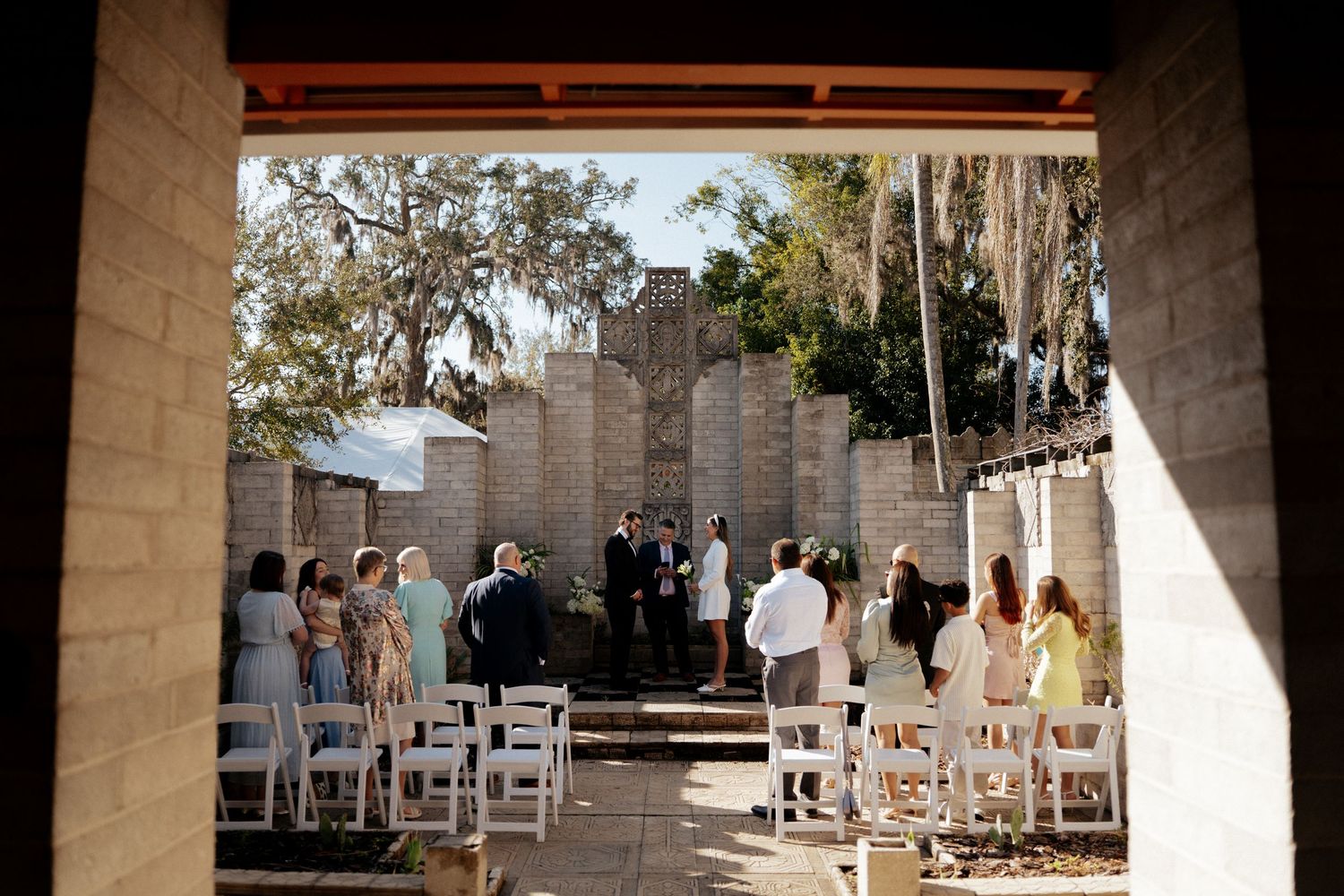 Outdoor wedding ceremony in a rustic courtyard with white chairs, stone walls, and lush trees in the background.