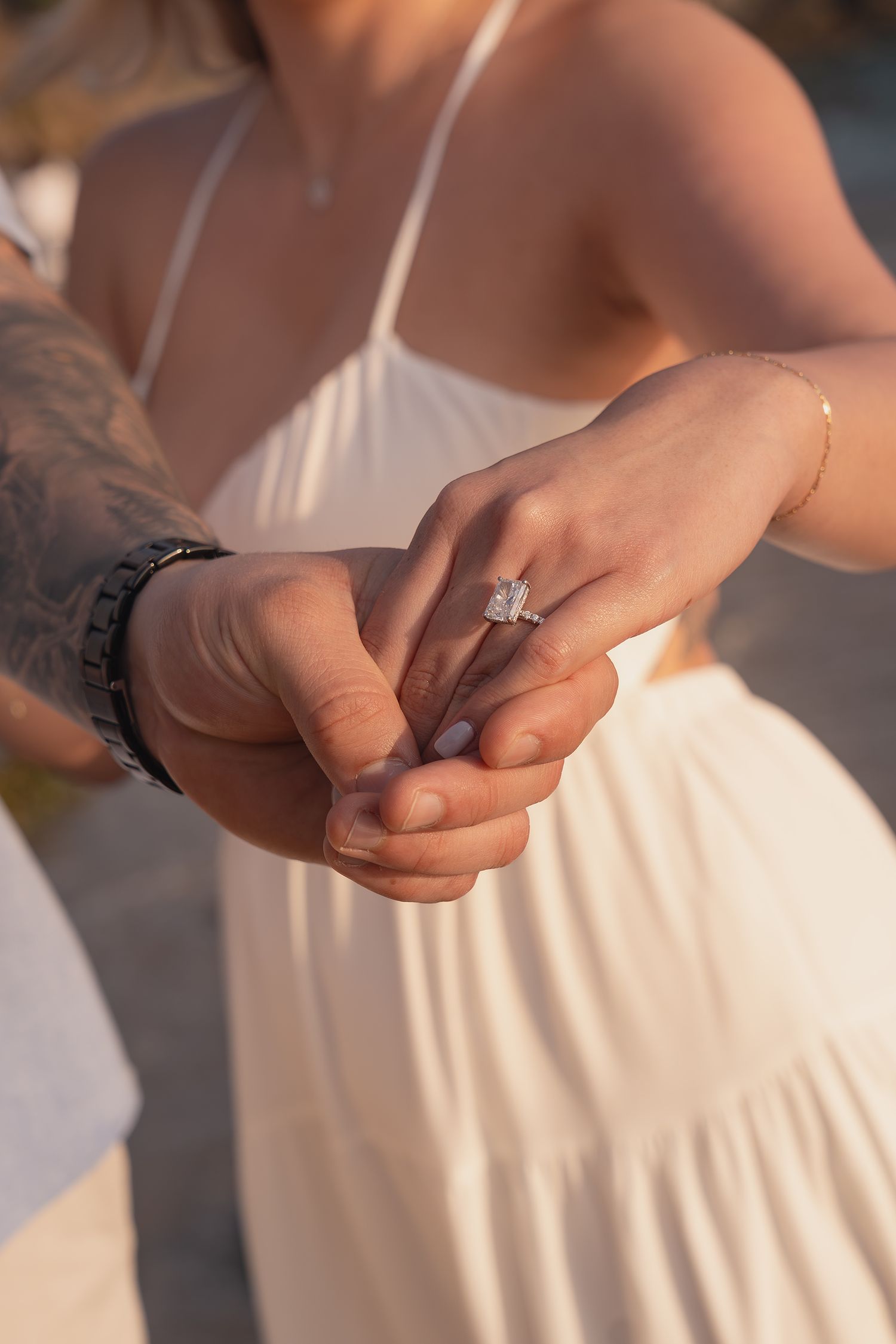 Close-up of couple holding hands showing a sparkling diamond engagement ring, woman wearing white dress.