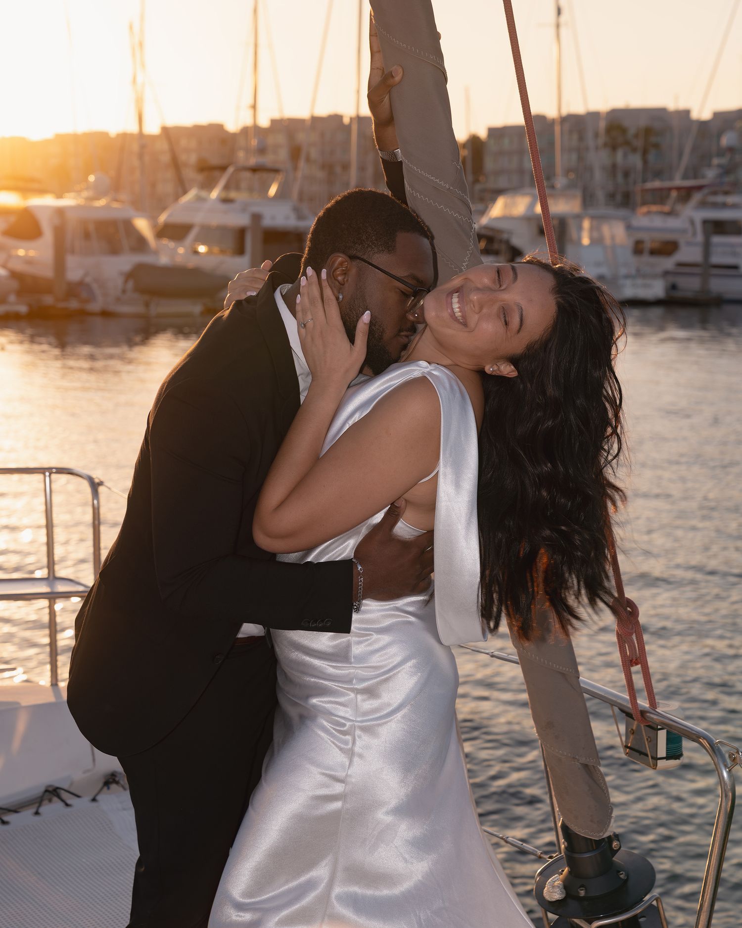 A couple embraces romantically on a sailboat at sunset in a marina.